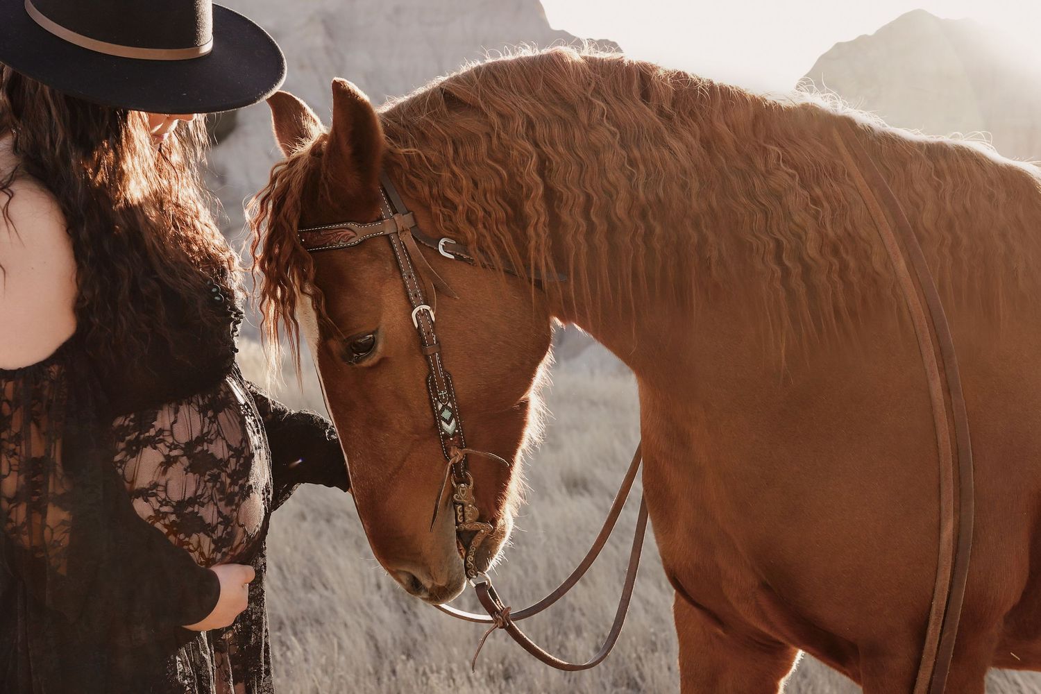 Pregnant woman in black lace dress gently touching chestnut horse outdoors.