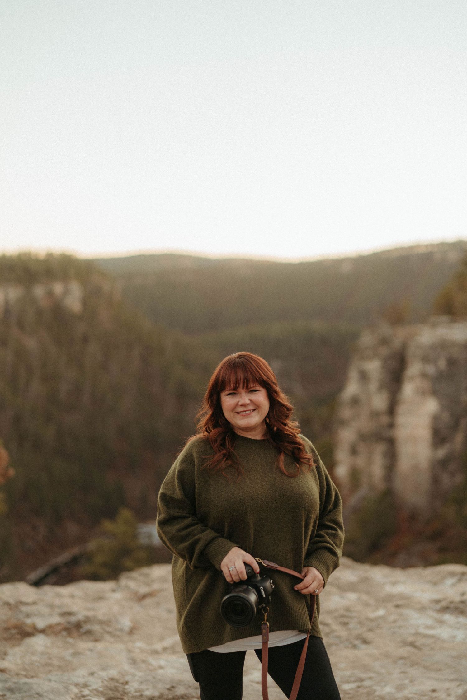 Smiling redhead female photographer holding DSLR camera standing on rocky overlook with mountain forest backdrop.