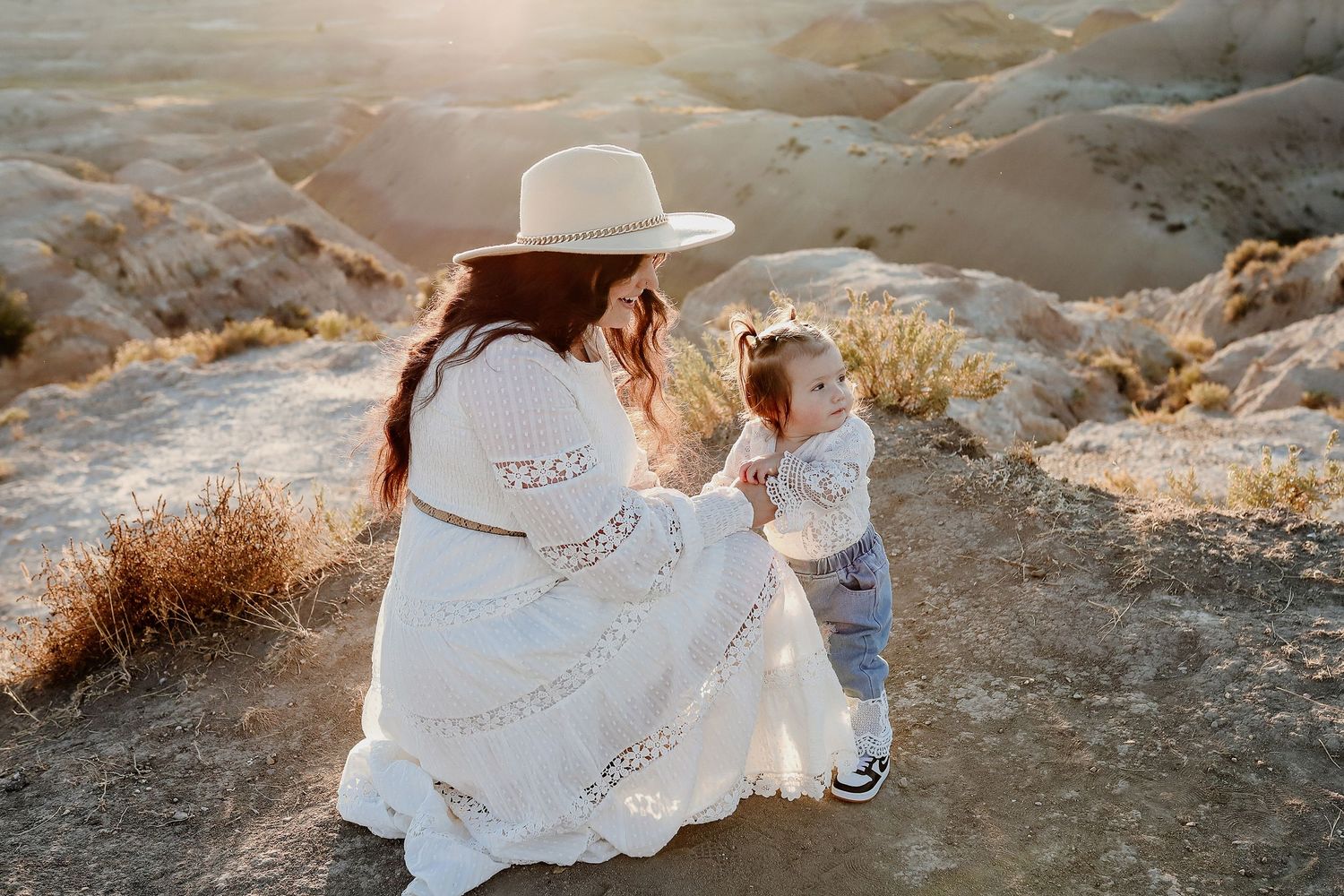 Mother in white boho dress and hat kneeling with toddler in a sunlit desert badlands landscape.