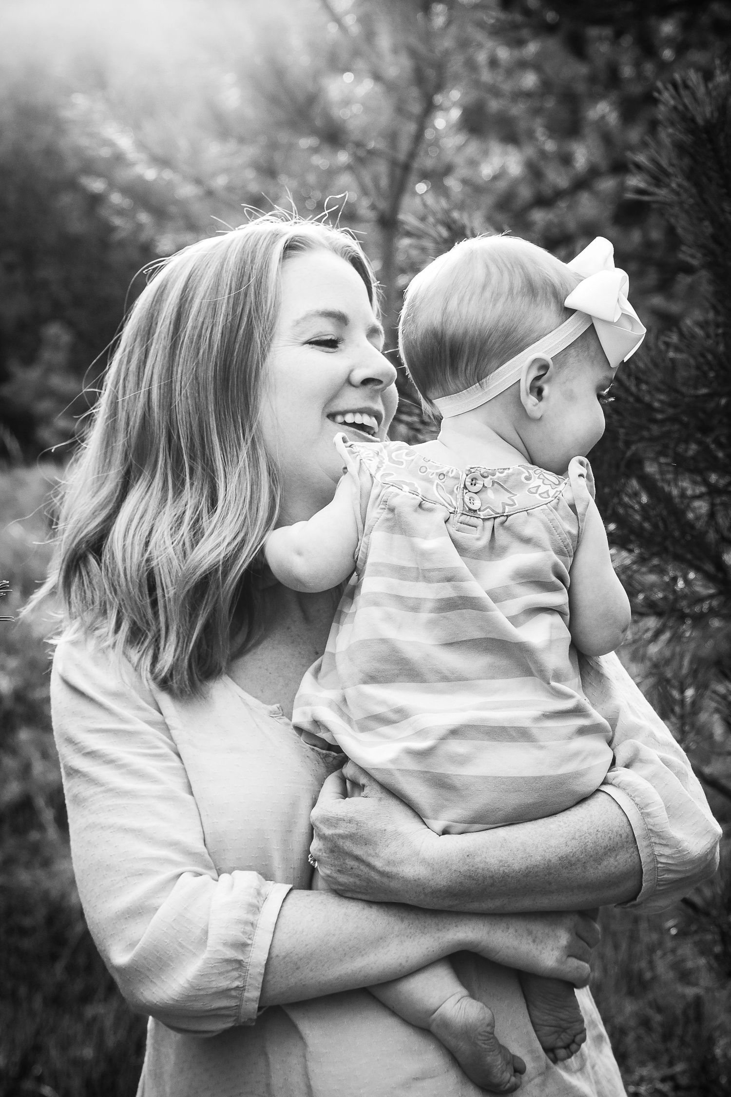 Laughing mother holds her baby girl outdoors near pine trees in black and white.