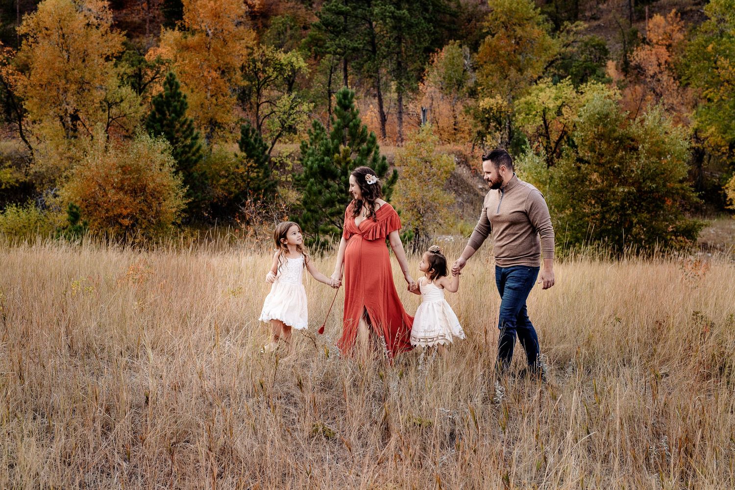 Family of four walks hand-in-hand through golden autumn meadow with colorful fall foliage.