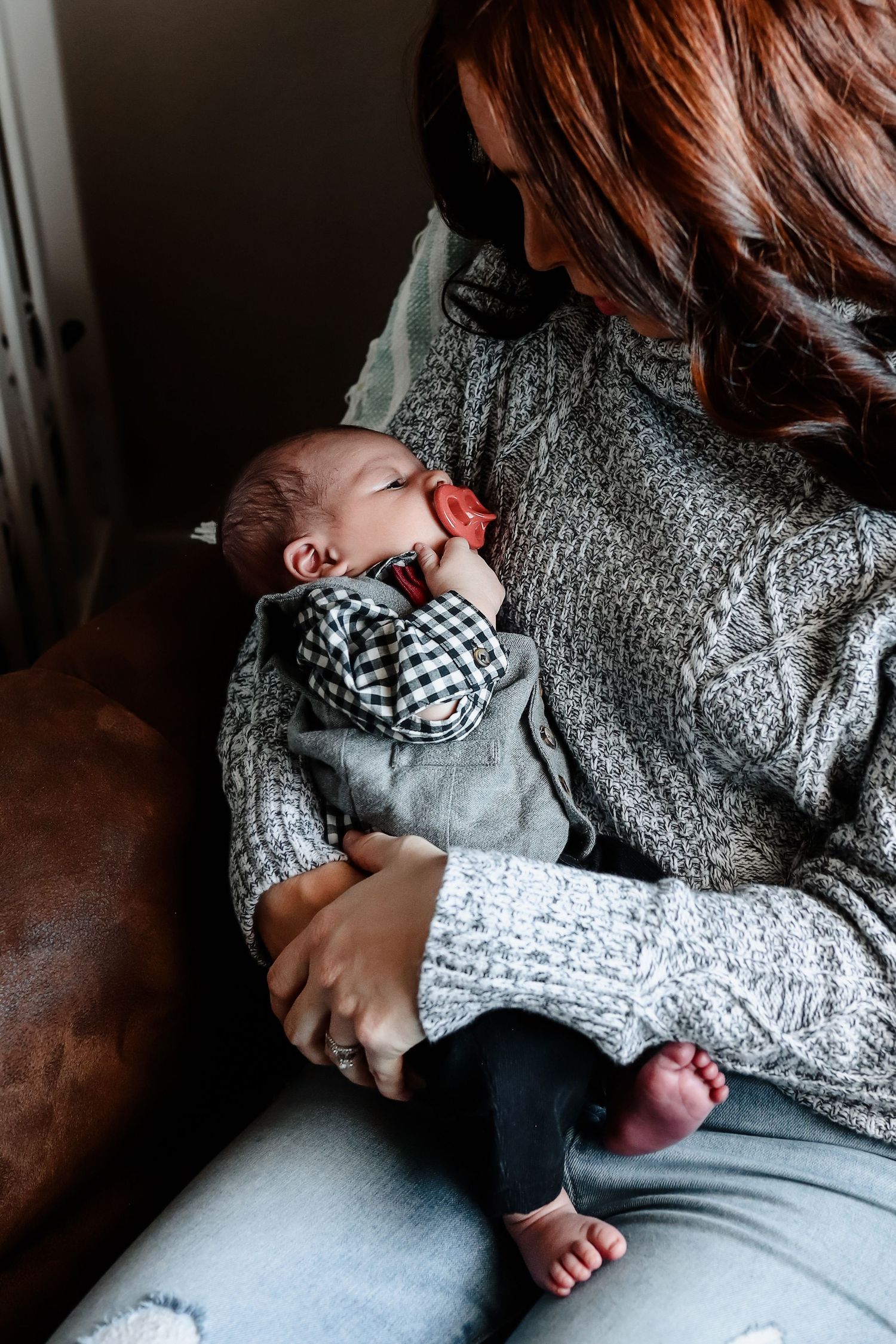 Close-up of newborn baby with pacifier sleeping peacefully in mothers arms wearing gray cable-knit sweater.