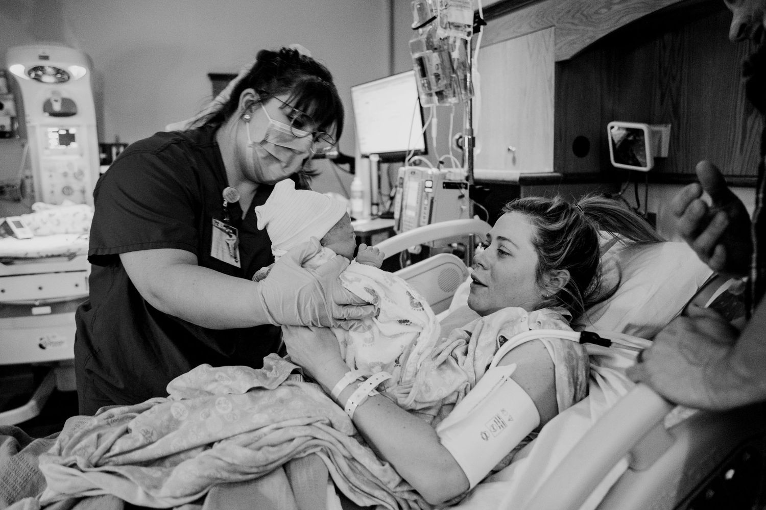 Nurse hands newborn baby to smiling mother in hospital labor and delivery room.