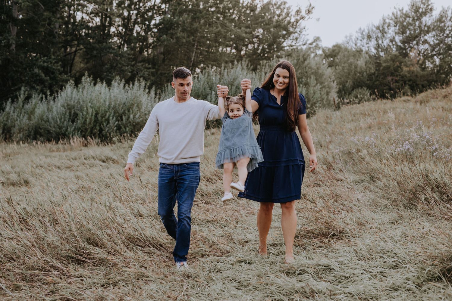 Happy family swinging toddler daughter while walking through a sunny meadow together.