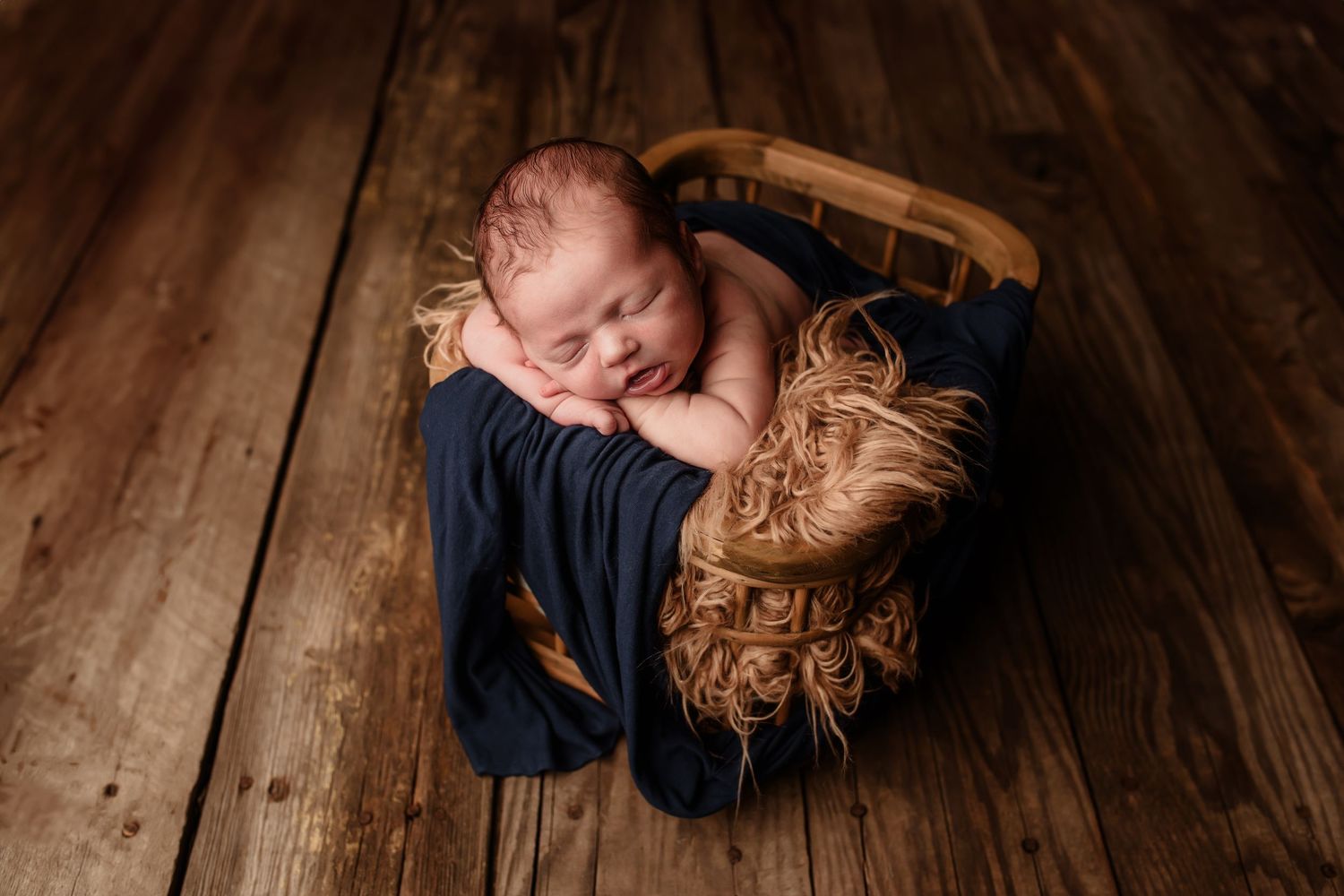 Sleeping newborn baby wrapped in navy blue cloth resting in a wooden bowl on rustic wood floor.