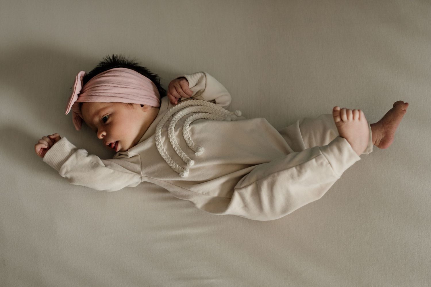 Peaceful newborn baby in cream striped outfit and pink headband sleeping on a soft sage green surface.