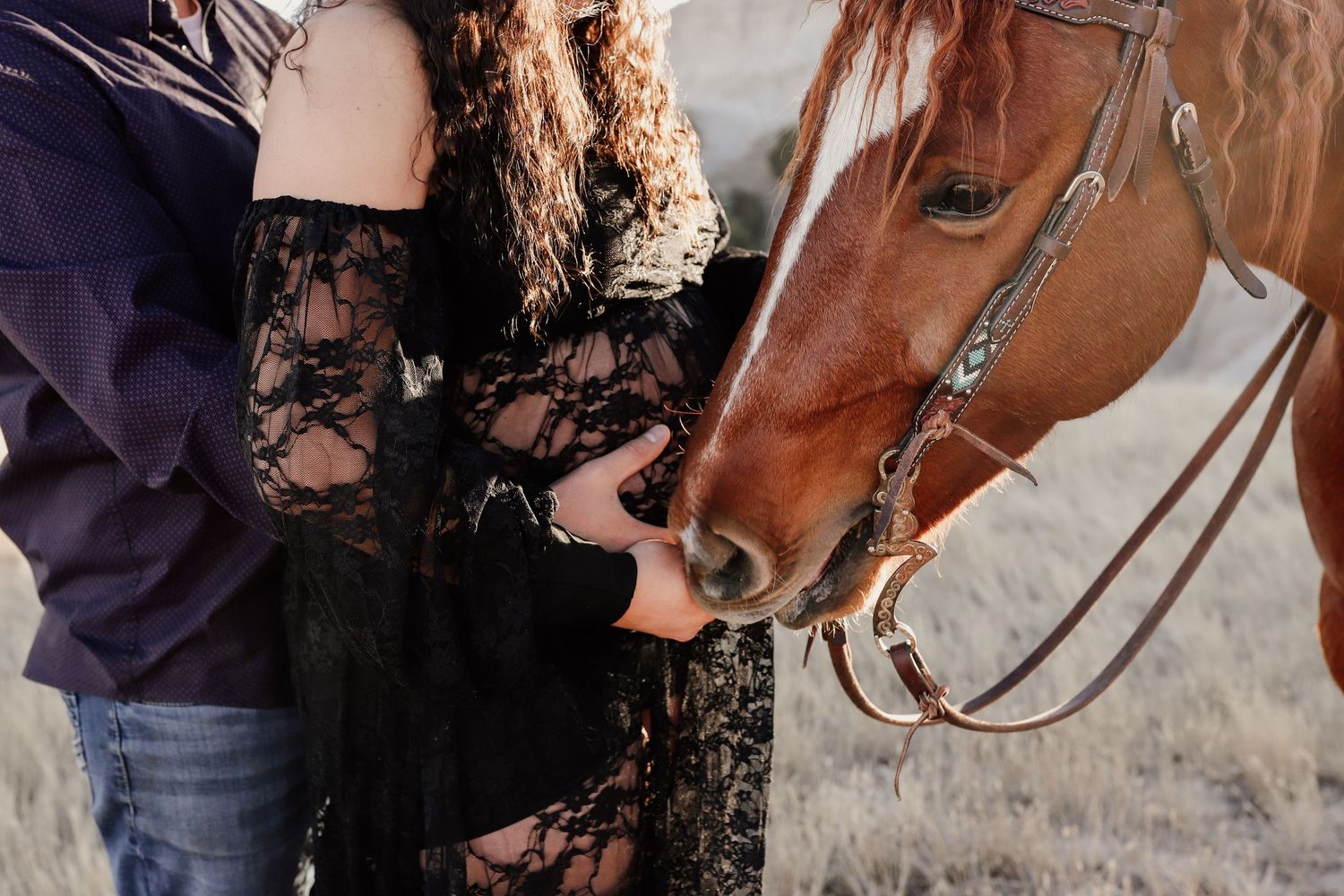 Couple in black lace and purple outfits feeding a chestnut horse up close in a dry field.
