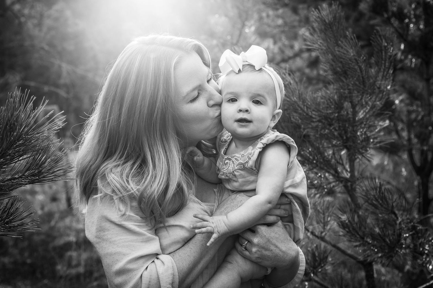 Mother gently kisses her baby daughters cheek outdoors among pine trees.