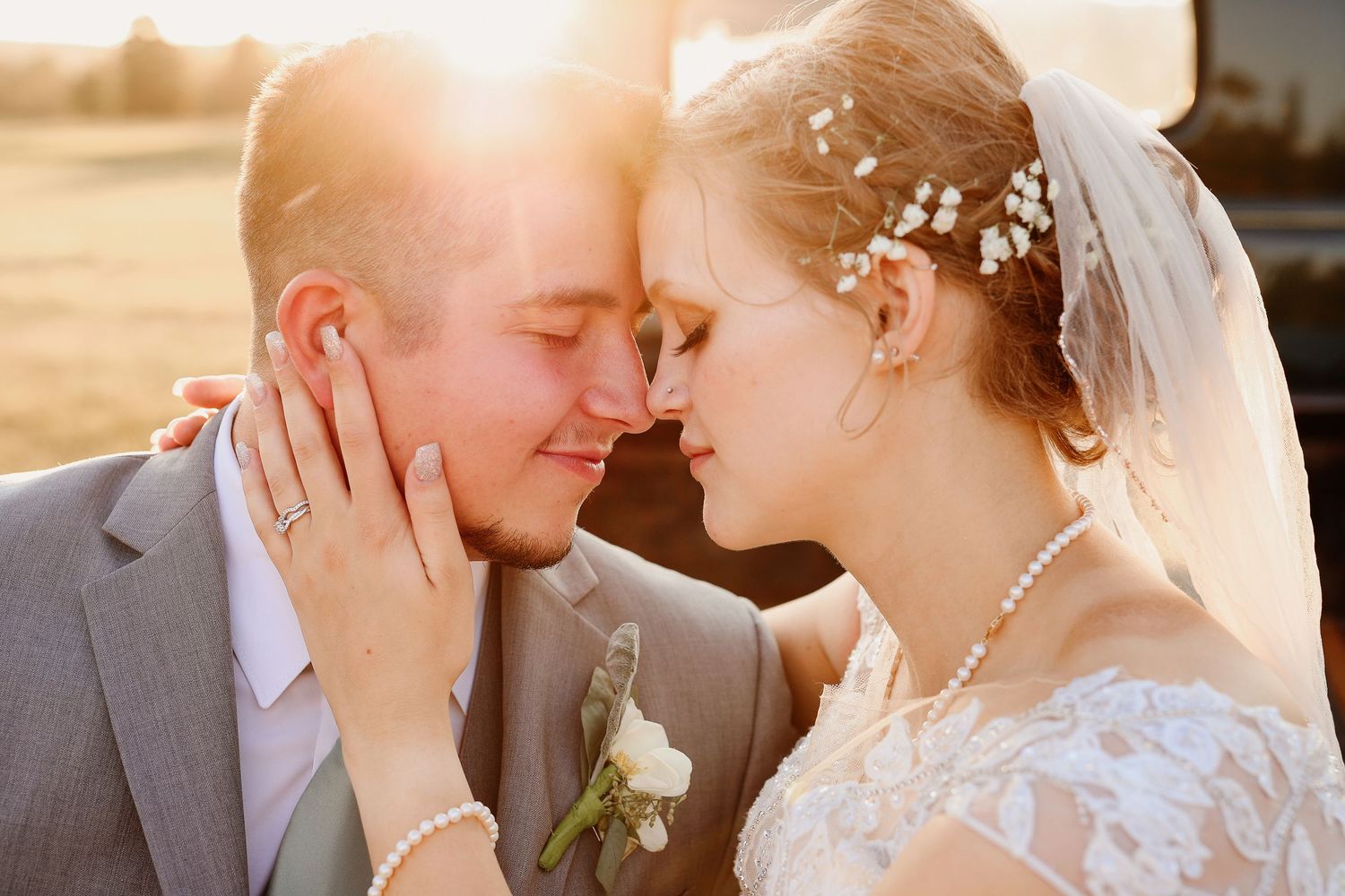 Bride and groom touching foreheads tenderly at golden hour sunset, bride wearing lace dress, veil, and pearl accessories.