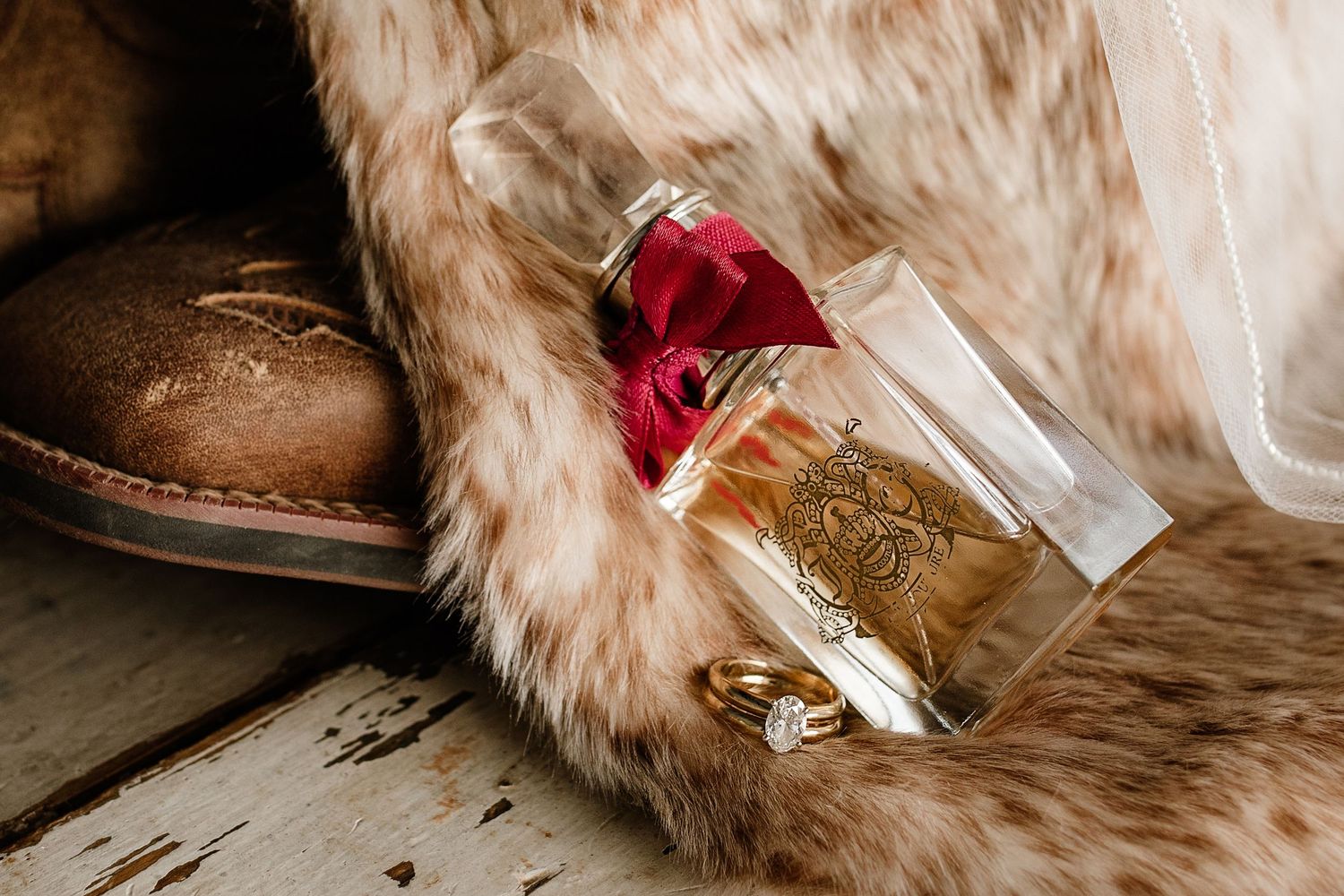 Luxury perfume bottle with red rose, gold wedding ring, and cowboy boot on rustic wood surface with fur accent.