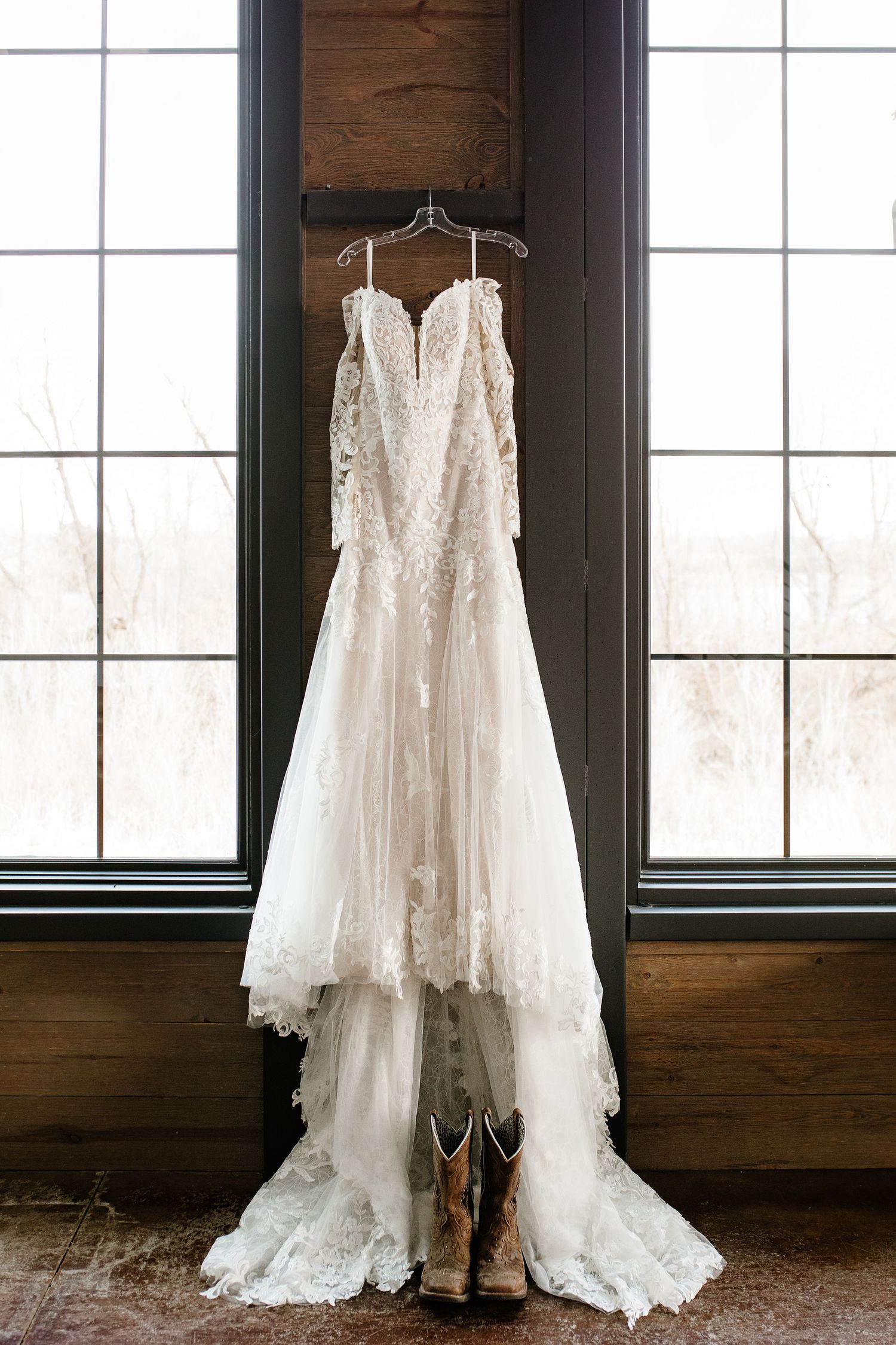 Lace wedding dress hanging by window with cowboy boots, elegant bridal gown with floral details and flowing skirt.