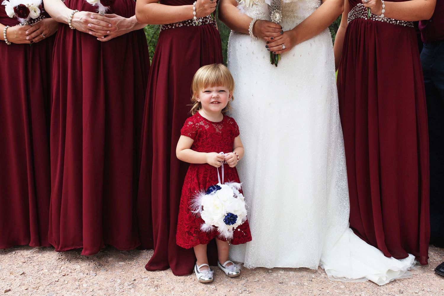 A young flower girl in a red dress holds a white bouquet, standing between bridesmaids in burgundy gowns and a bride.