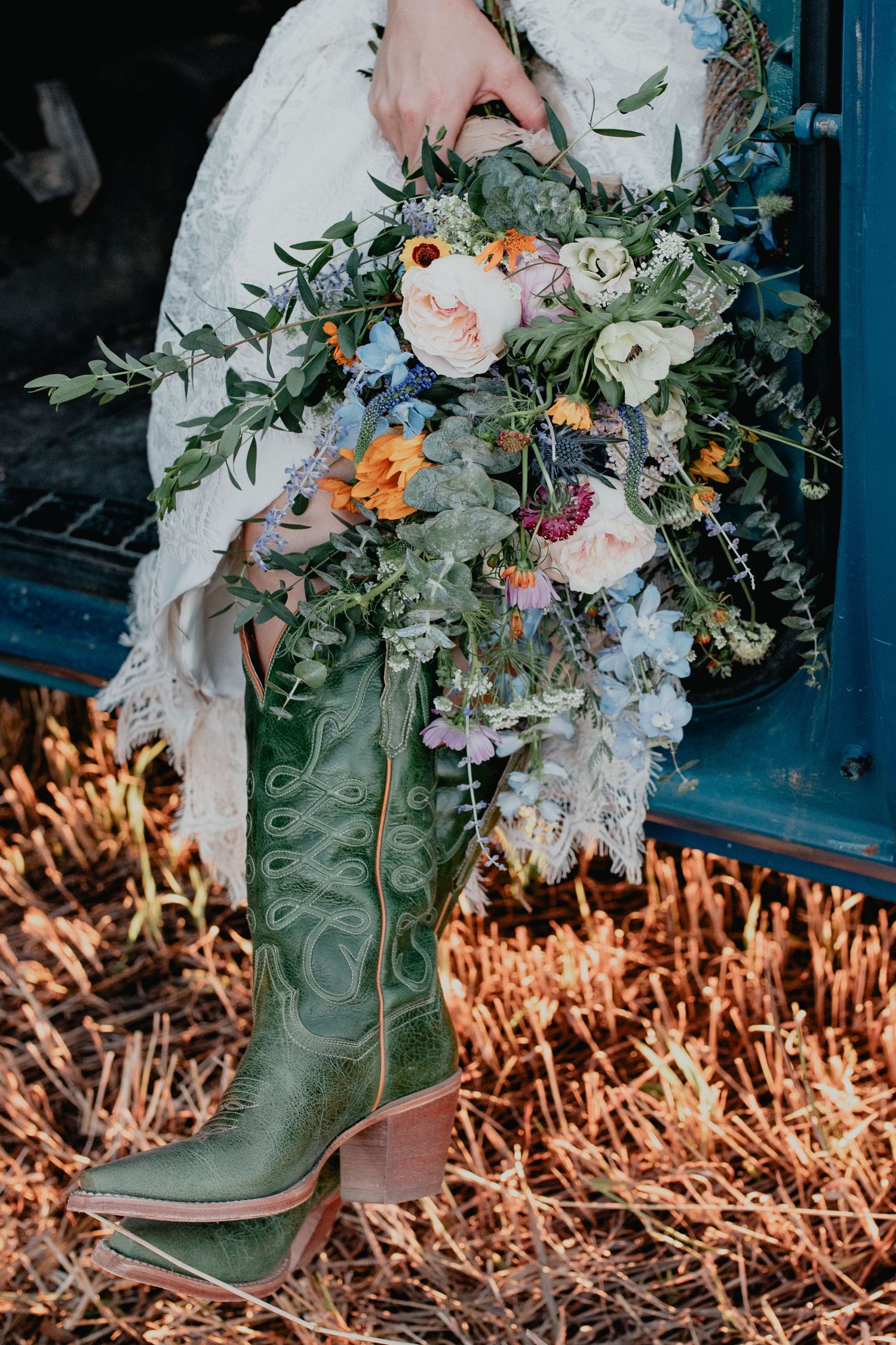 Bride in lace dress holding wildflower bouquet wearing green cowboy boots, sitting on vintage truck tailgate.