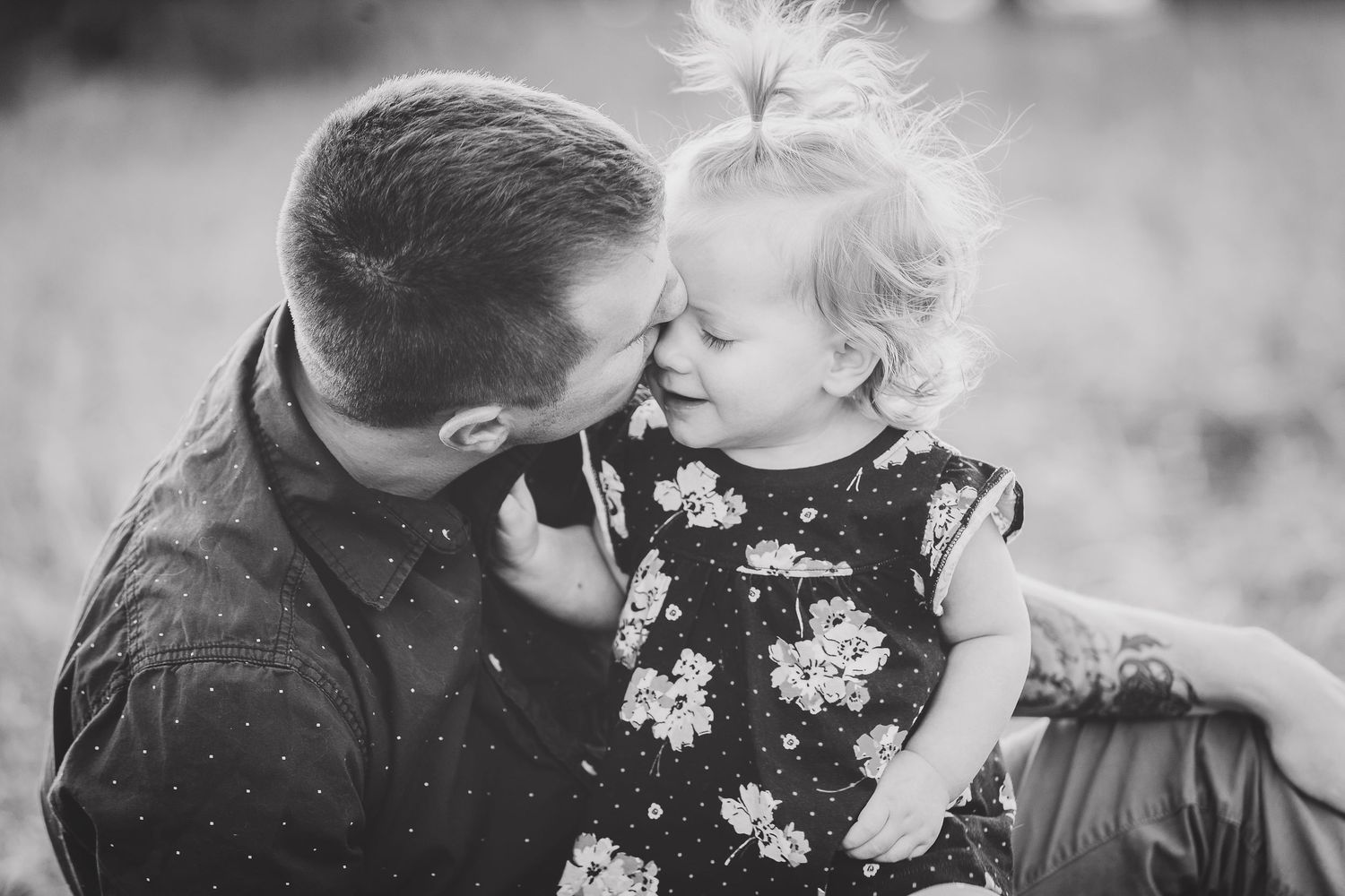 Father affectionately nuzzling his smiling toddler daughter in a field.