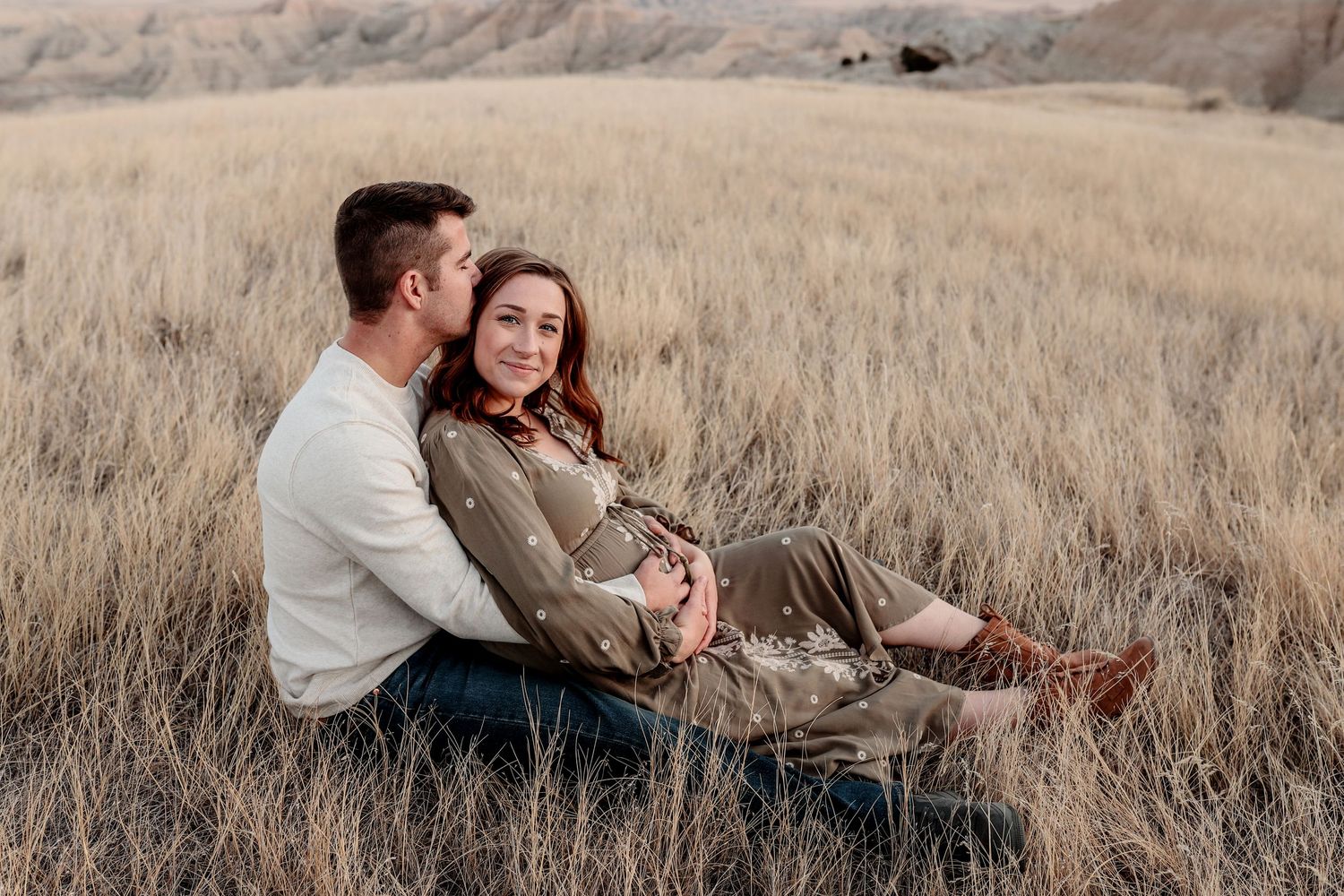 Expecting couple sitting together in golden dry grass field with badlands backdrop.
