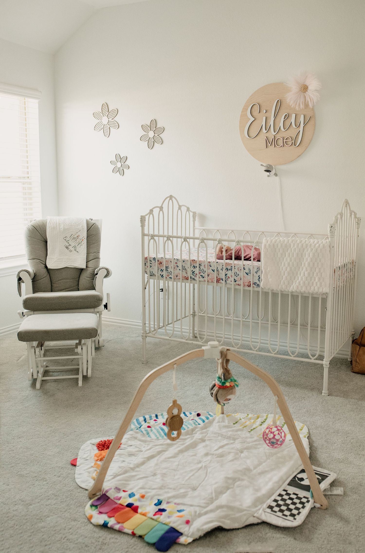 Bright neutral nursery with white iron crib, gray glider, colorful play mat, and personalized name sign for Eiley.