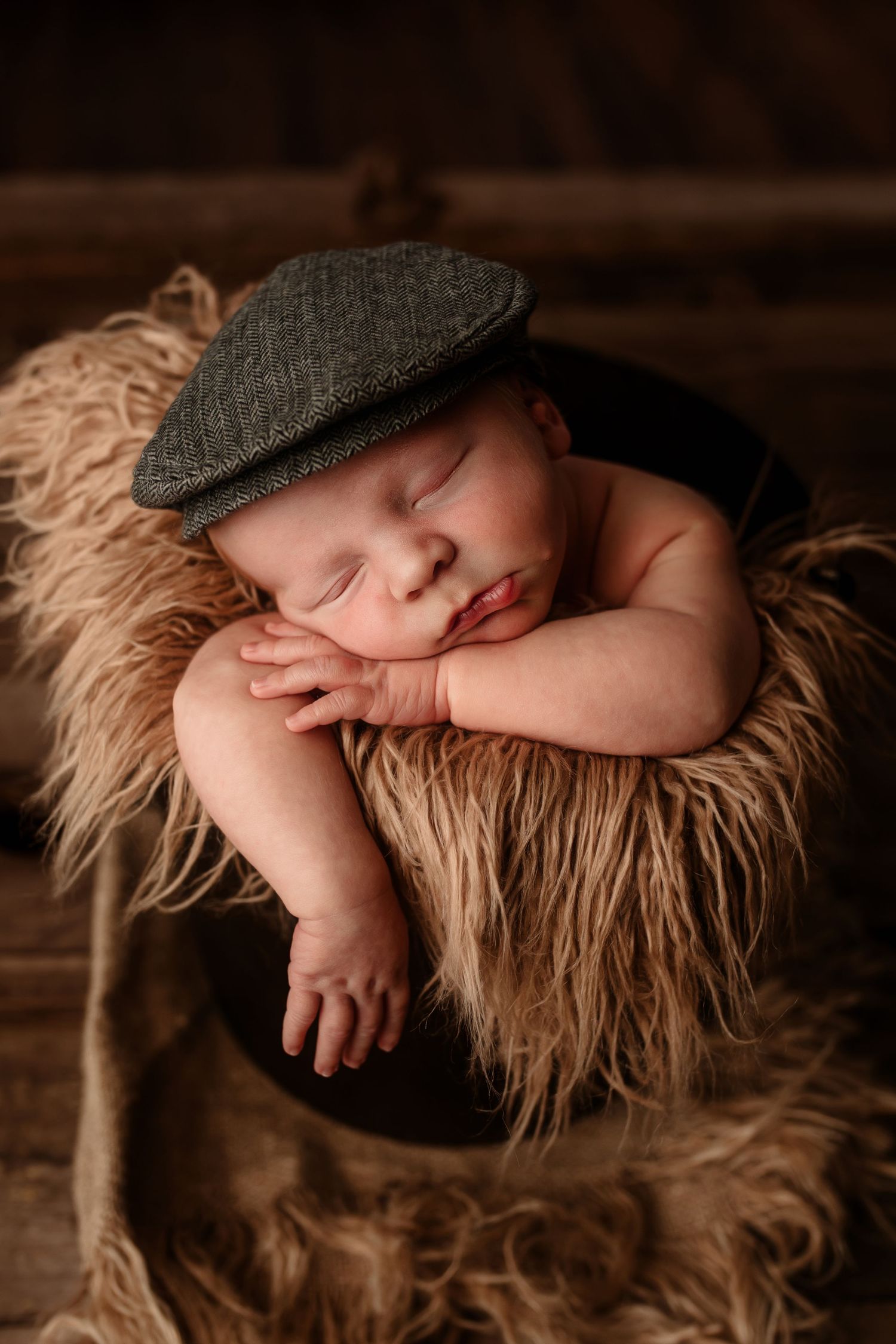 Sleeping newborn baby boy wearing a gray flat cap hat posed in a fuzzy brown bucket.