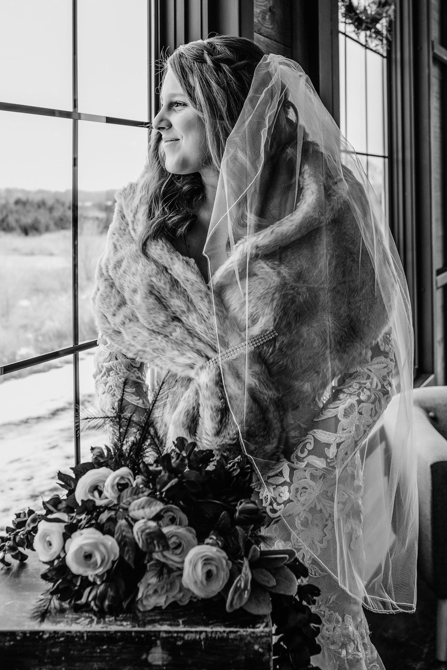 Black and white photo of a bride in lace veil and fur wrap gazing out a window, holding a bouquet of roses.