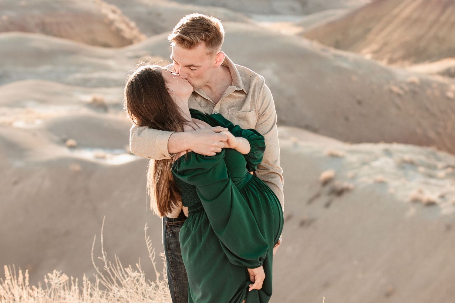 Two people share a romantic embrace while standing in a scenic mountain landscape at sunset.
