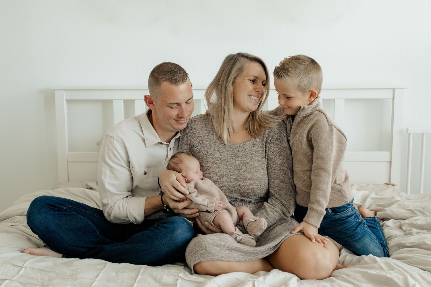 Happy family of four with mother, father, newborn baby, and young son sitting together on white bed.