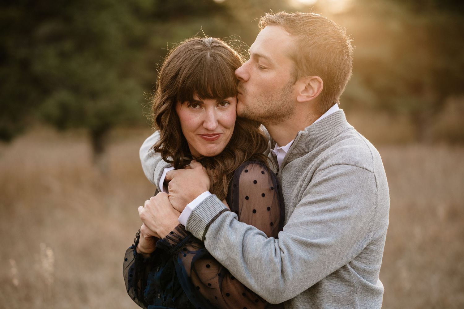 Couple embracing at golden hour in a field, man kissing woman's cheek in a romantic outdoor portrait.