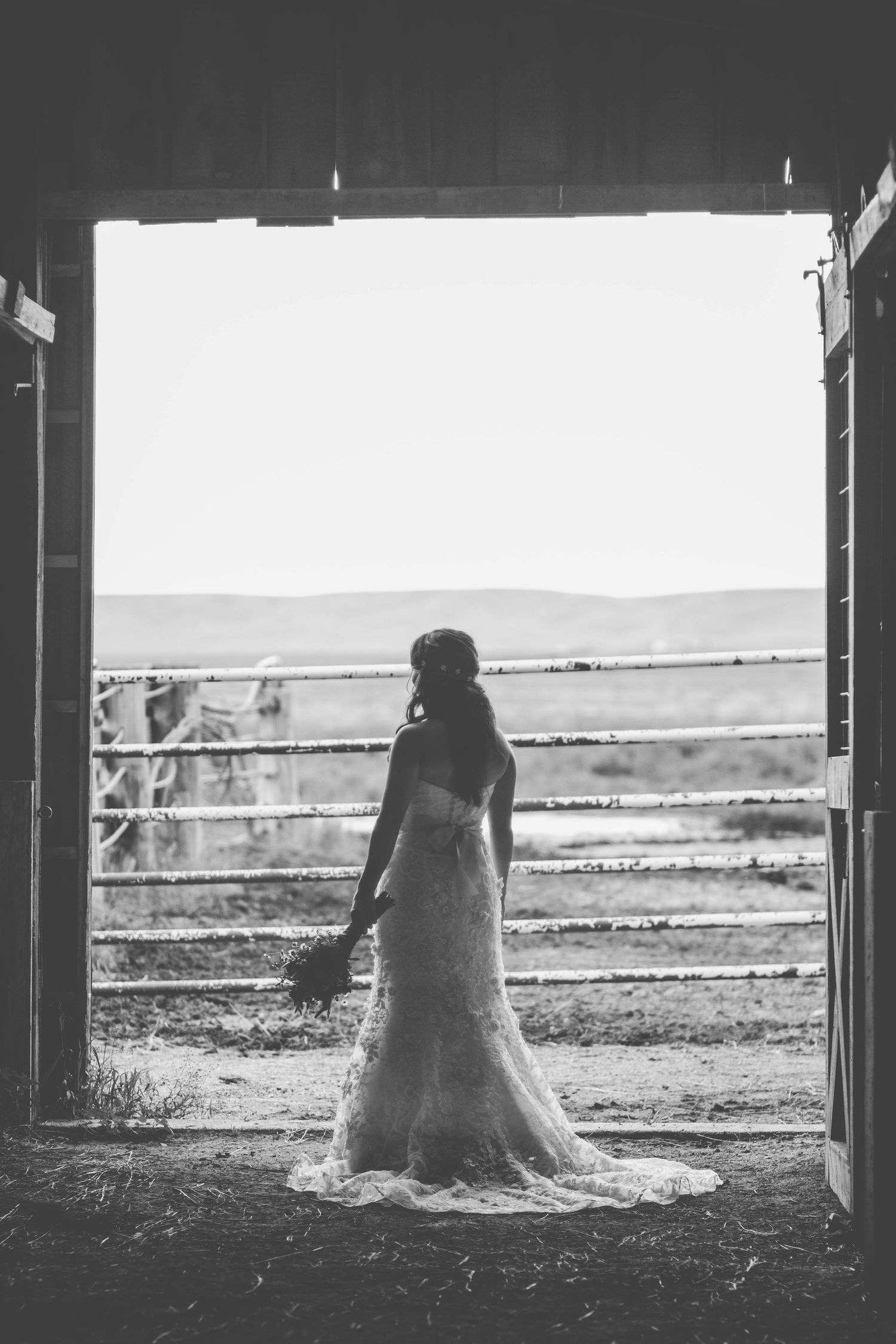 Black and white photo of a bride in a lace gown standing in a barn doorway holding a bouquet.