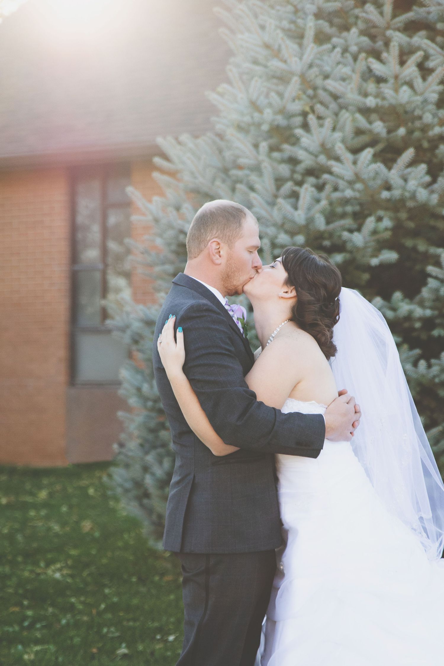 Bride and groom share a romantic kiss outdoors near an evergreen tree, bride in white gown with veil, groom in dark suit.