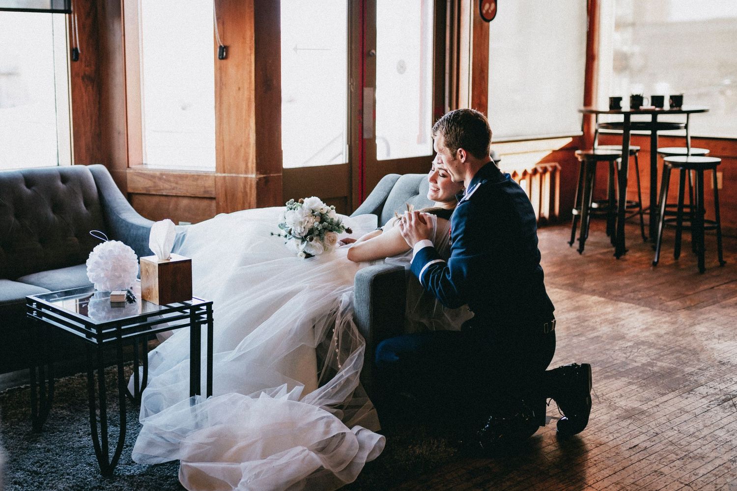 Groom kneeling before bride in elegant wedding venue, sharing an intimate moment beside a floral bouquet.