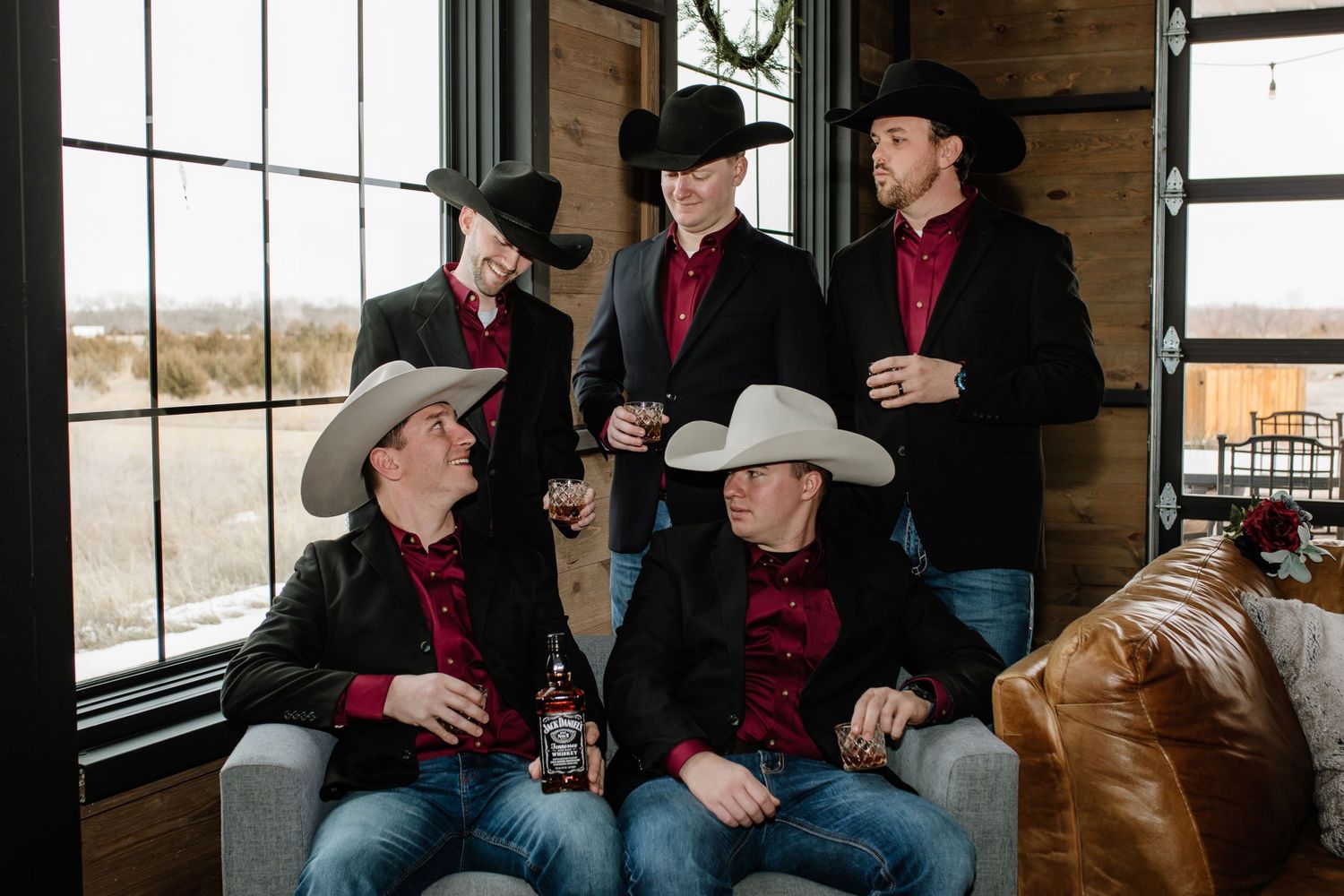 Five men in black blazers and cowboy hats pose together, holding drinks in a rustic wood-paneled venue.
