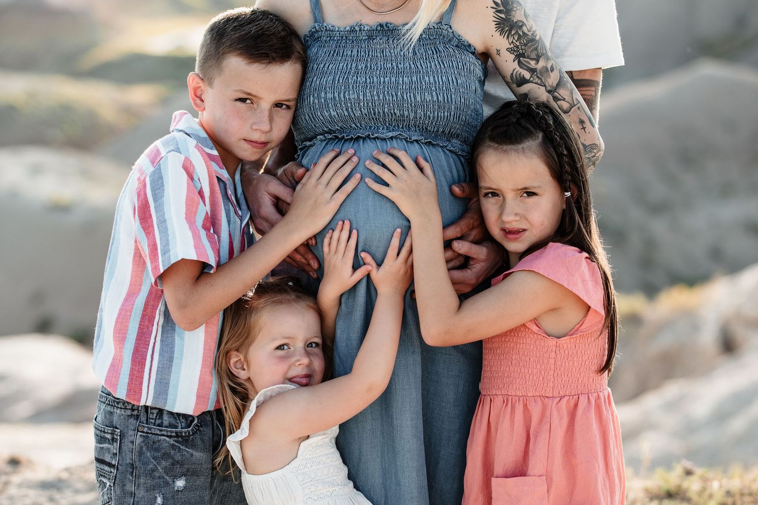 Three children lovingly embrace their pregnant mother's baby bump outdoors during a maternity photo session.