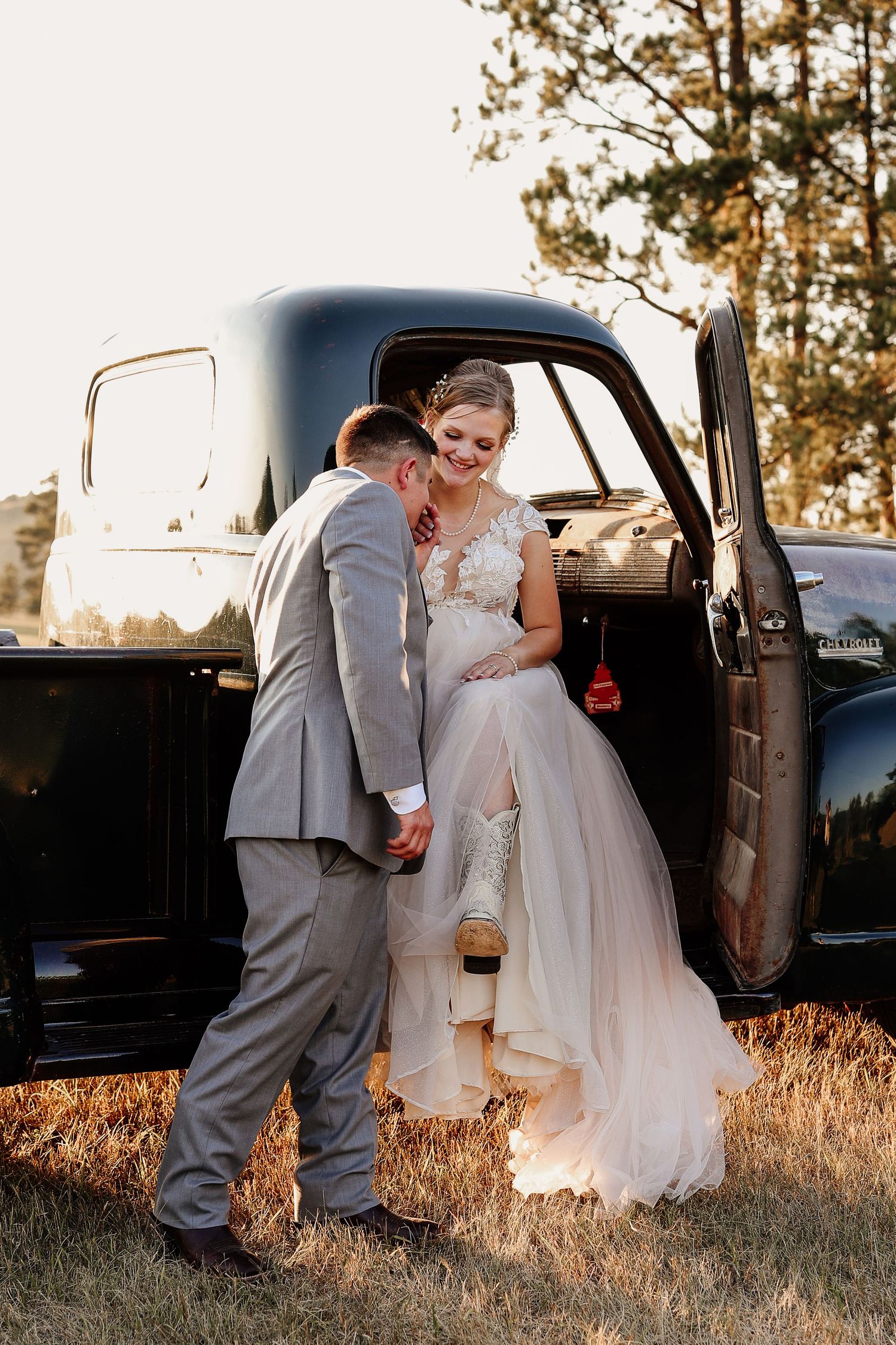 Bride in white gown and cowboy boots sits in vintage black truck as groom in gray suit assists her at golden hour.