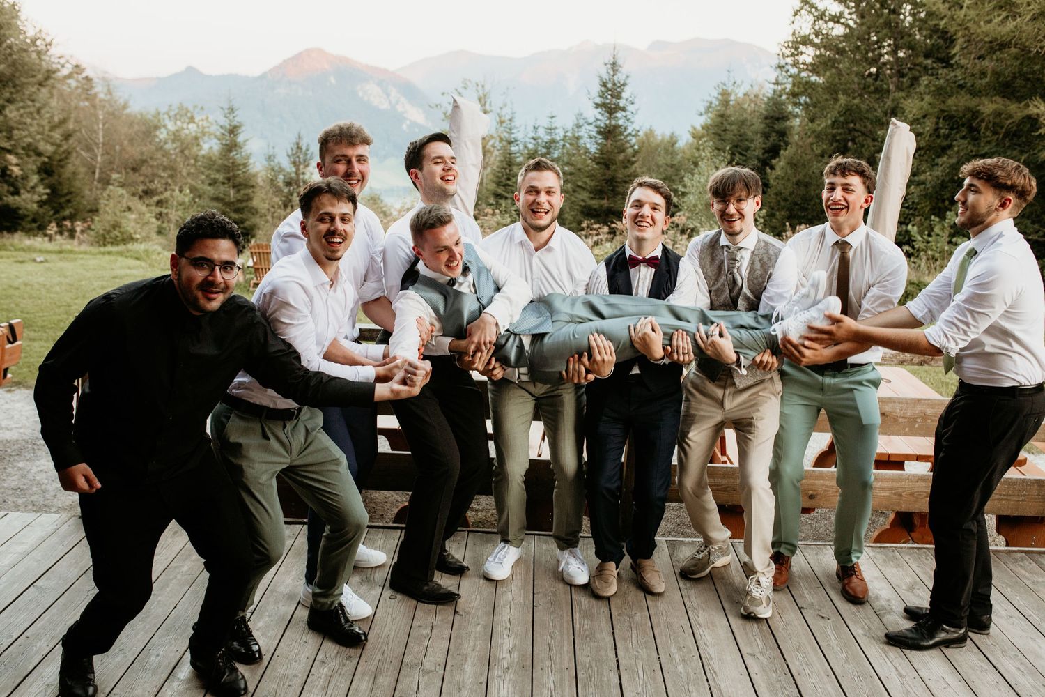 Groomsmen lifting groom horizontally outdoors at mountain wedding venue, all smiling in white shirts.