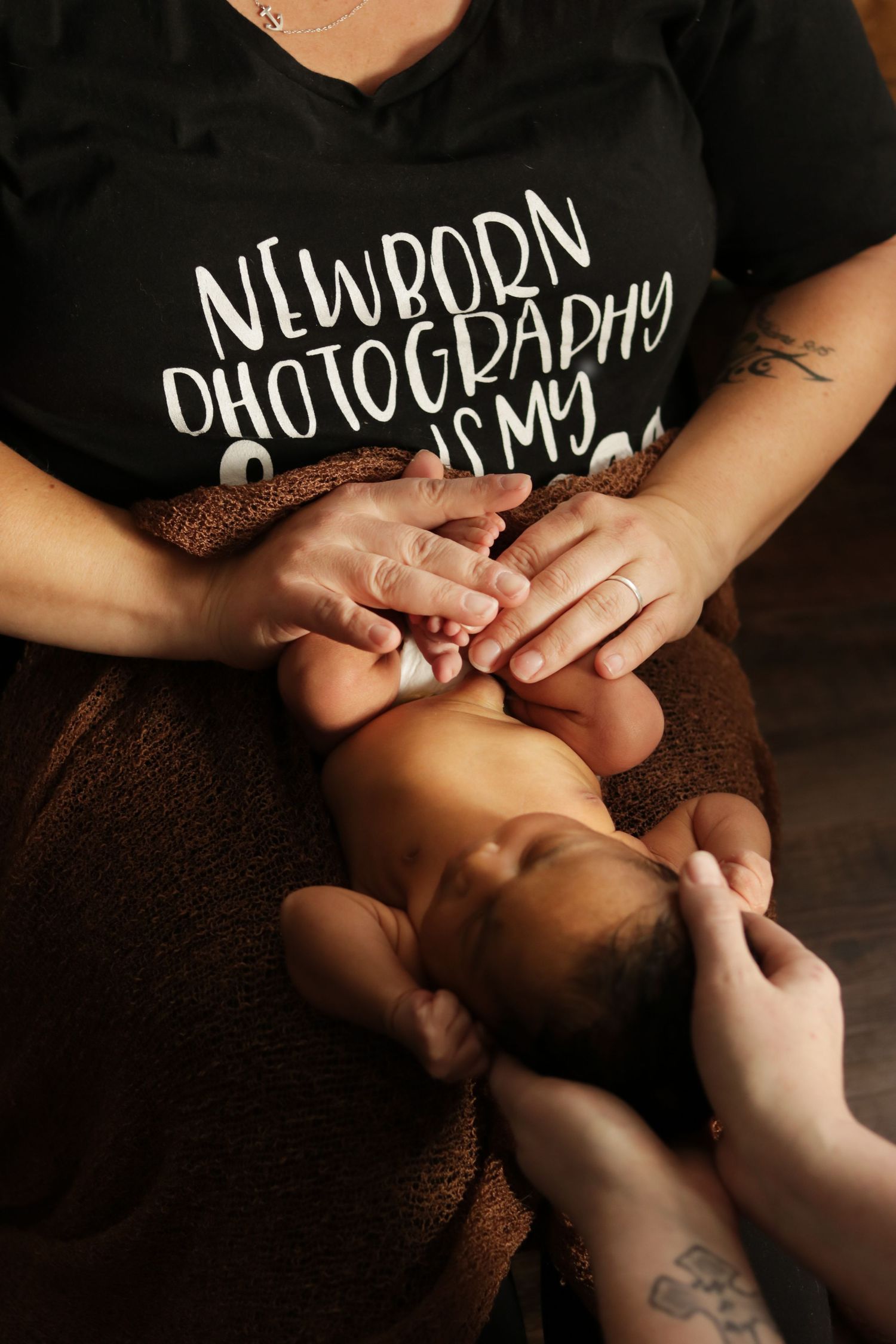 Photographer wearing a 'Newborn Photography' t-shirt holds a tiny baby on a brown blanket during a photo session.