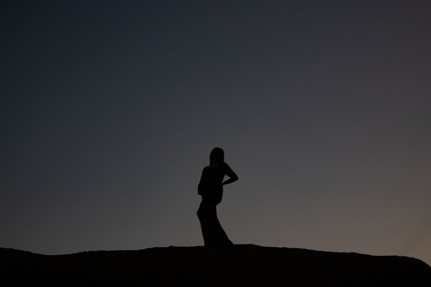 Silhouette of pregnant woman standing against a deep blue dusk sky at sunset.