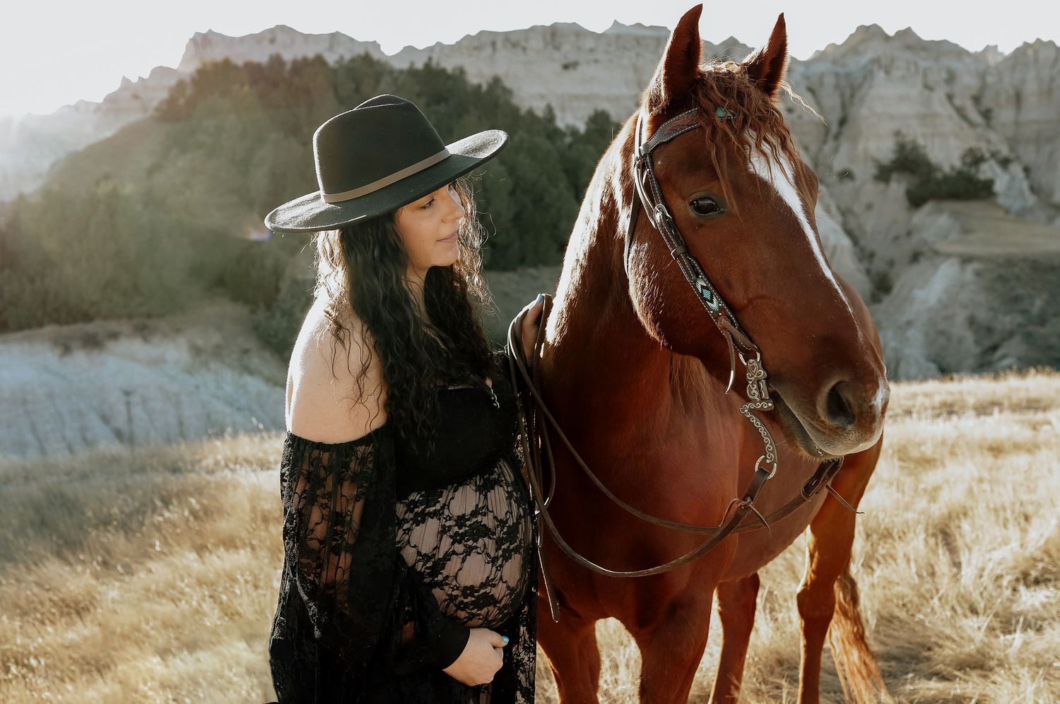 Pregnant woman in black lace dress and wide-brim hat standing beside chestnut horse in scenic badlands landscape.