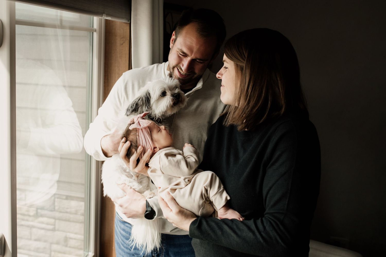 Happy parents laughing by a window as their fluffy white dog sniffs their sleeping newborn baby.