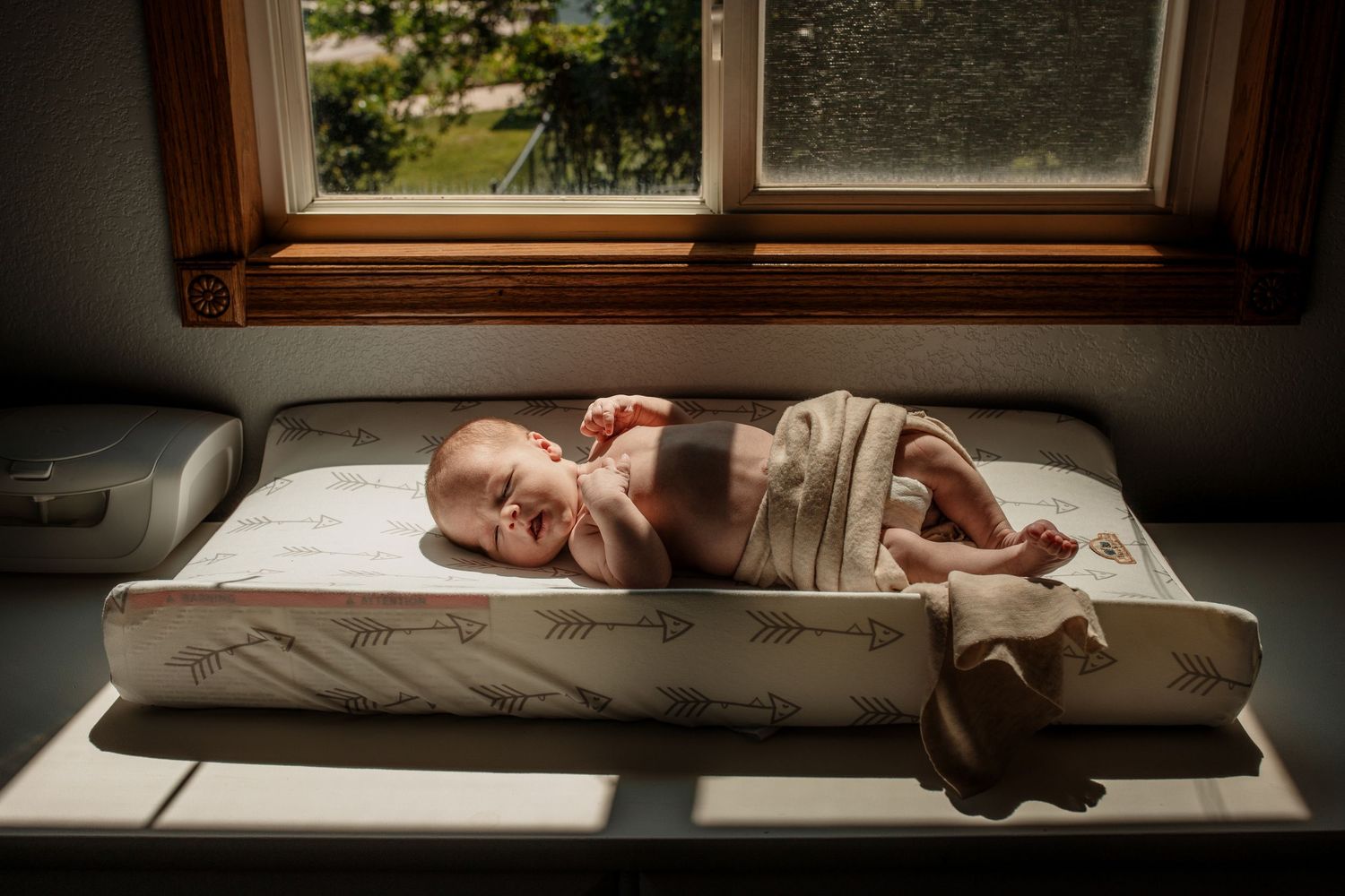 Newborn baby sleeping peacefully on a changing pad in warm sunlight by a window.