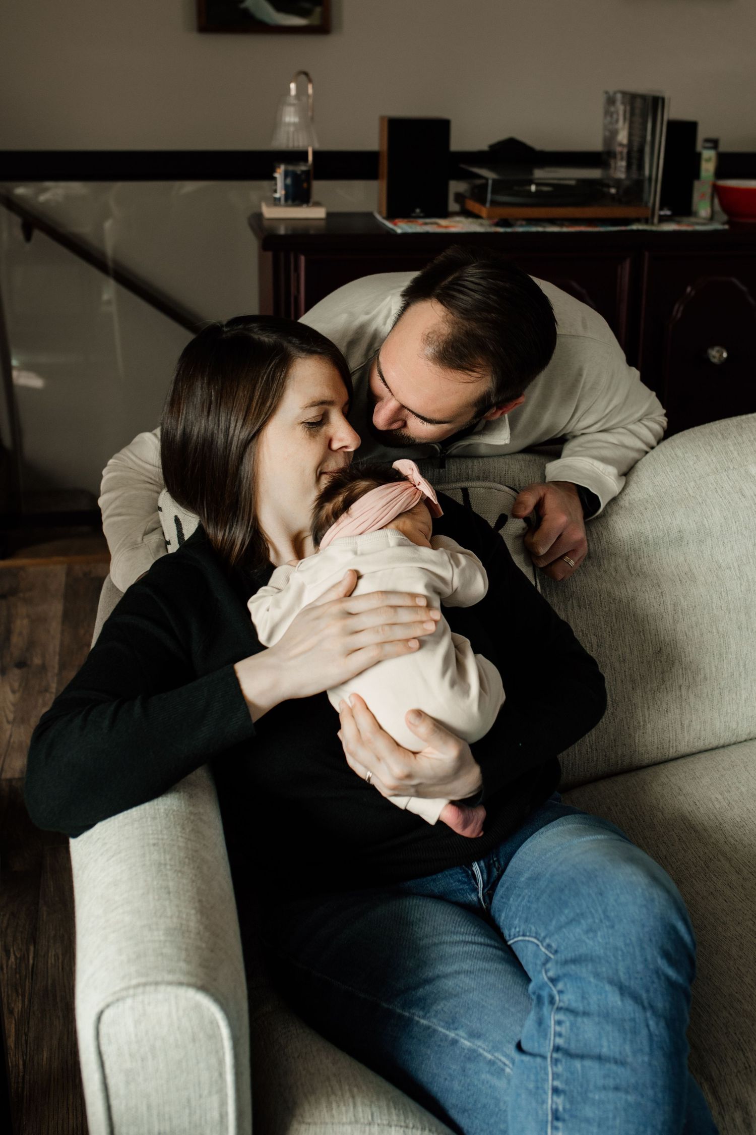 Parents share a tender moment kissing their newborn baby together while seated in a cozy armchair.