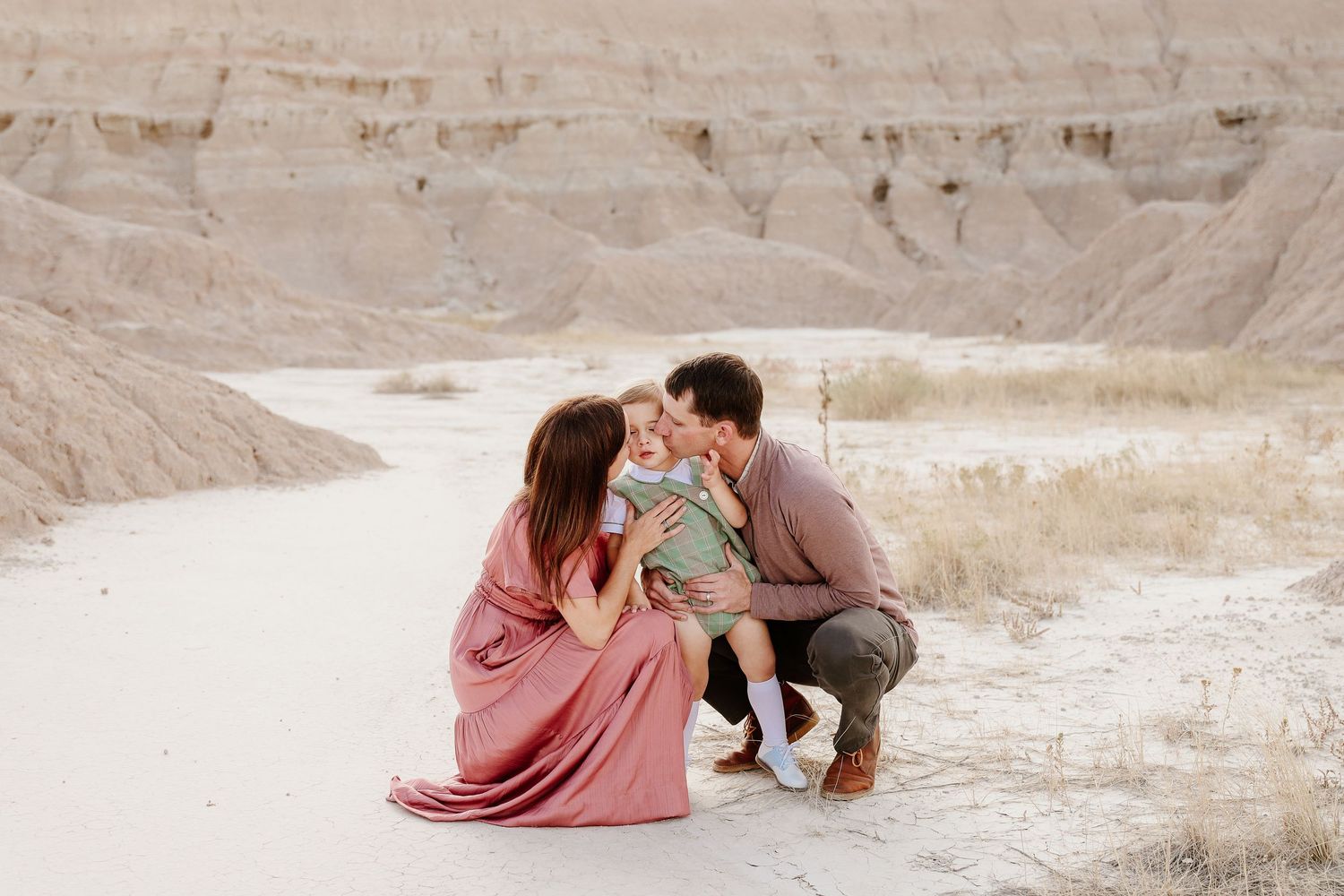 Family of three in desert landscape, parents kissing toddler in green outfit, mother in pink dress.