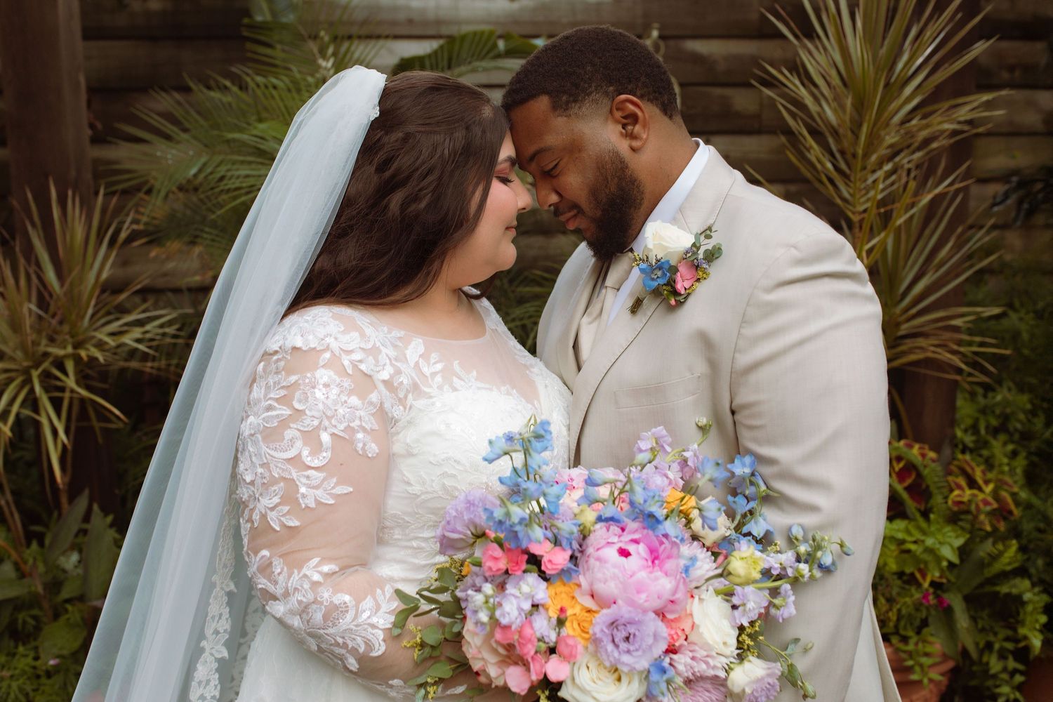 Newlywed couple in wedding attire touching foreheads, bride holding colorful pastel bouquet with pink peonies and blue flowers.