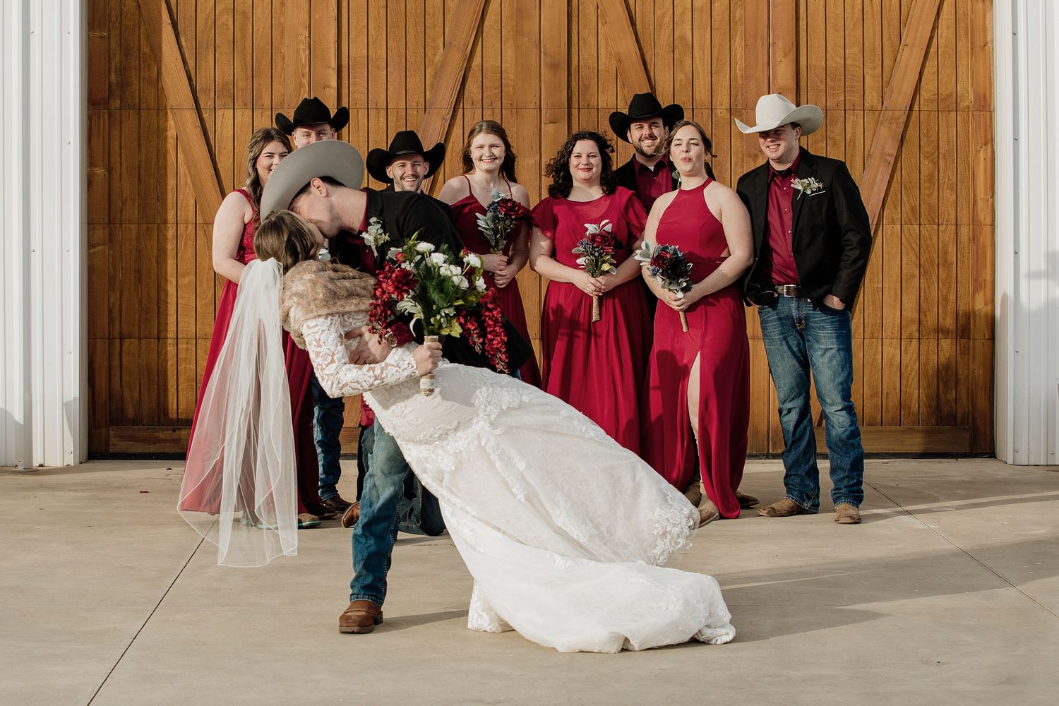 Bride and groom share a dip kiss at a rustic barn wedding with bridesmaids in red dresses and groomsmen in cowboy hats.