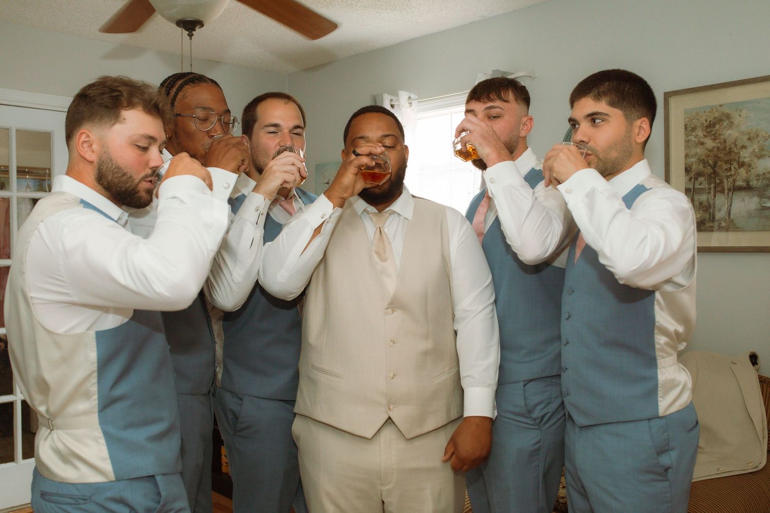 Groom in cream vest with groomsmen in blue suits taking a celebratory shot before the wedding ceremony.