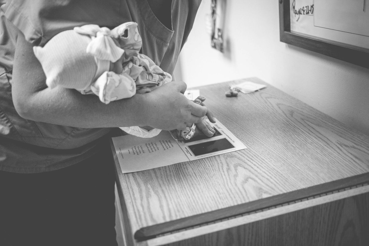 Parent holding a swaddled newborn baby while standing at a wooden hospital bedside table.
