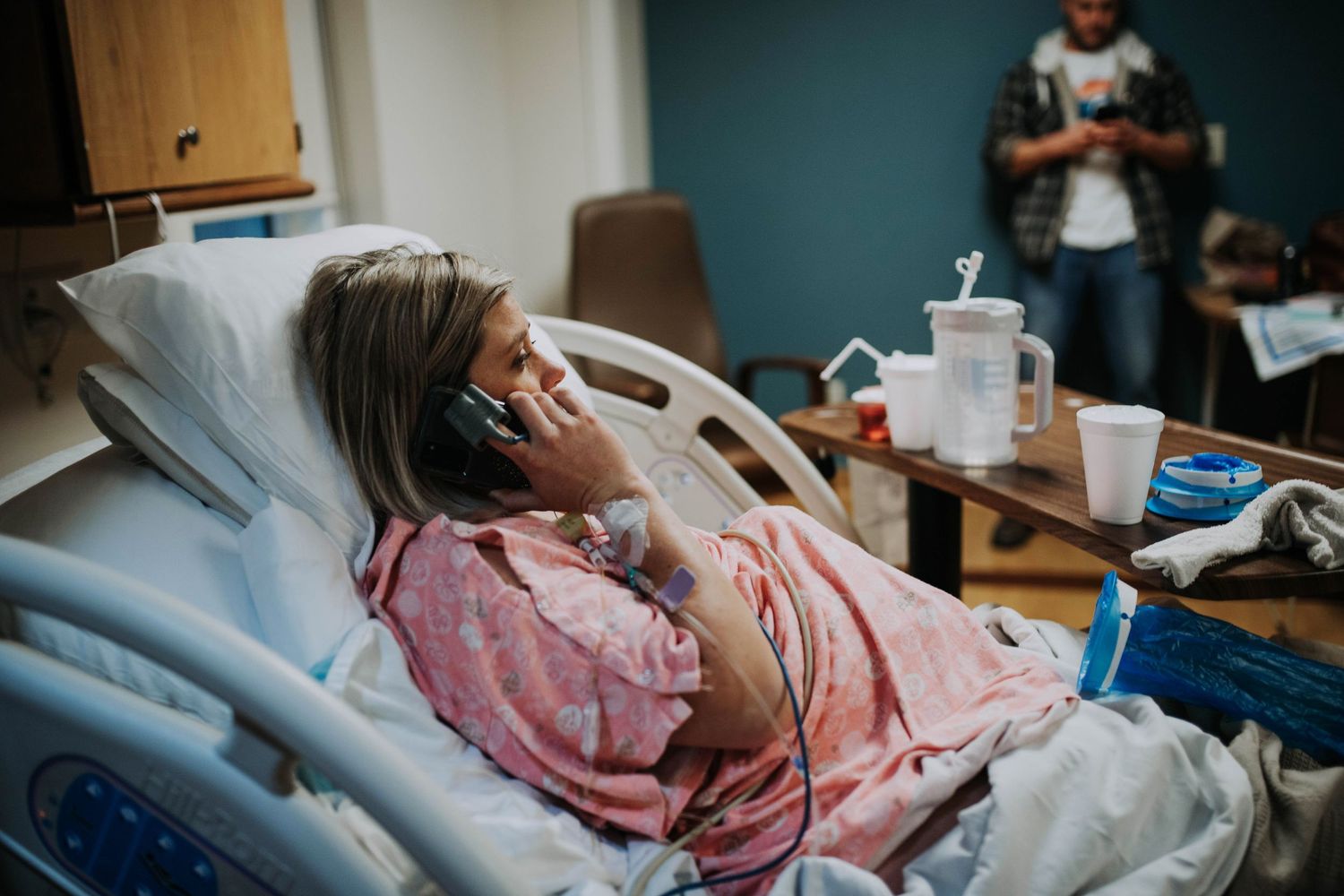 Pregnant woman lying in hospital bed talking on phone while partner stands in background.