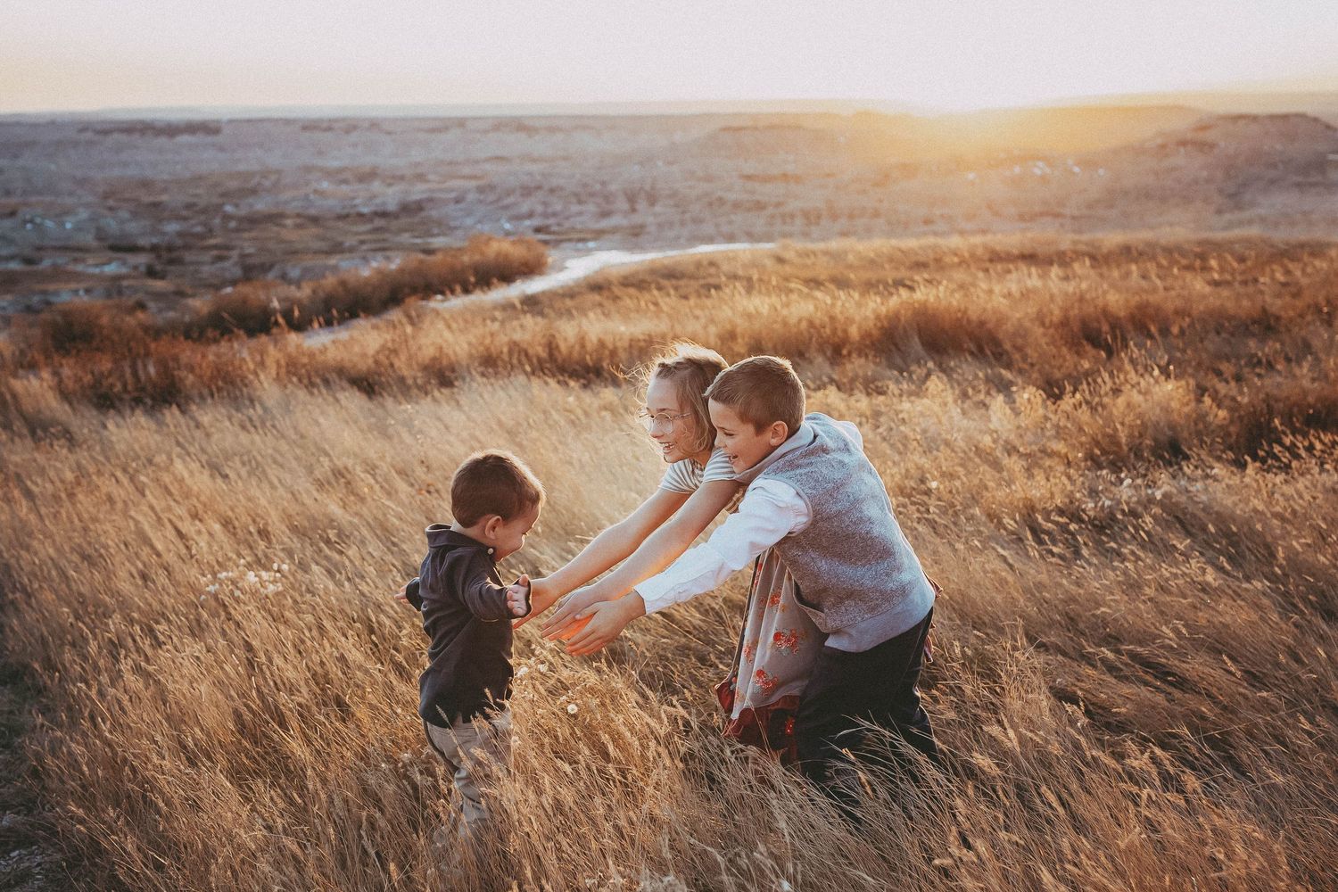 Parents kneeling in golden grass field at sunset, engaging lovingly with their young child.