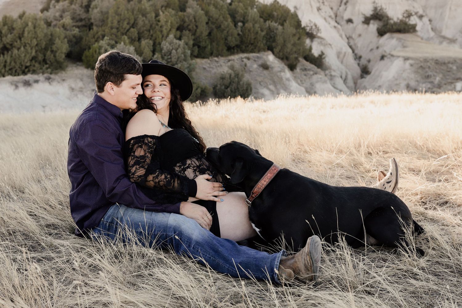 Couple sitting in golden grass field with black Labrador dog, woman in black lace dress wearing wide-brim hat.