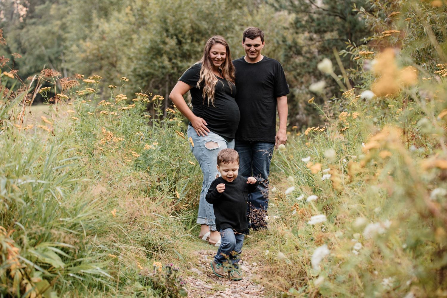 Pregnant woman and man in black shirts smile with toddler on a wildflower trail outdoors.