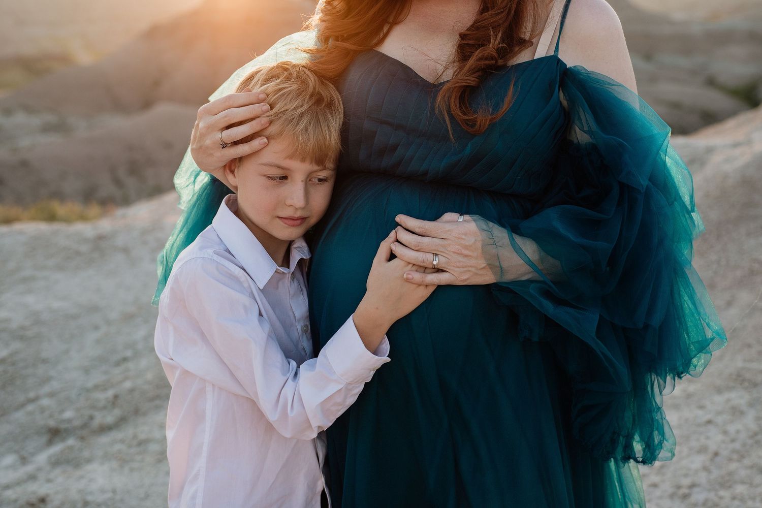 Young boy hugs pregnant woman in teal gown at sunset in rocky desert landscape.