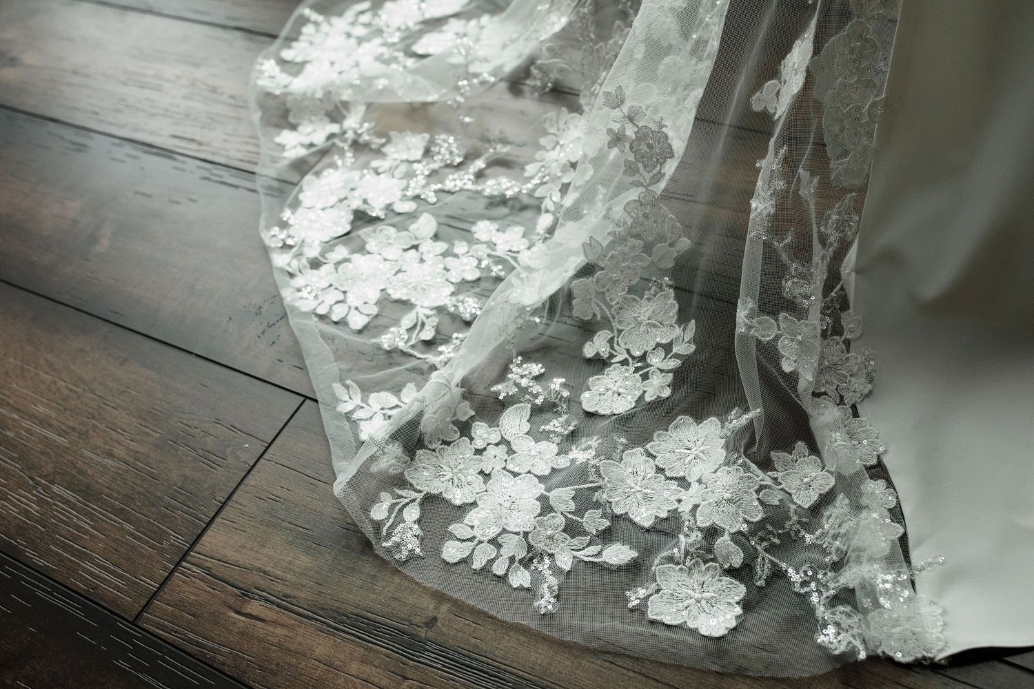 Close-up of elegant white floral lace wedding dress hem on dark hardwood floor.