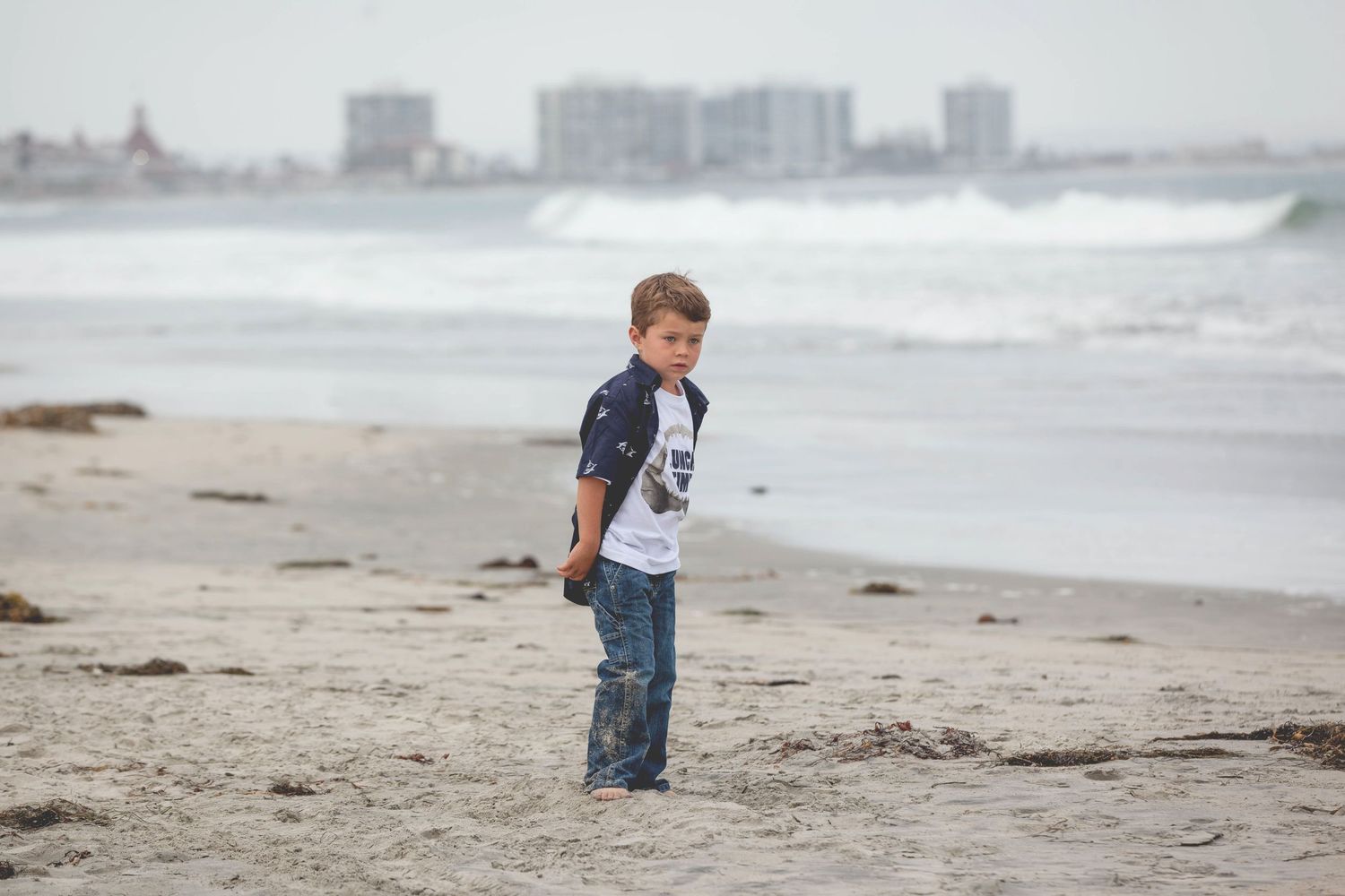 Young boy stands alone on a sandy beach with ocean waves behind him.